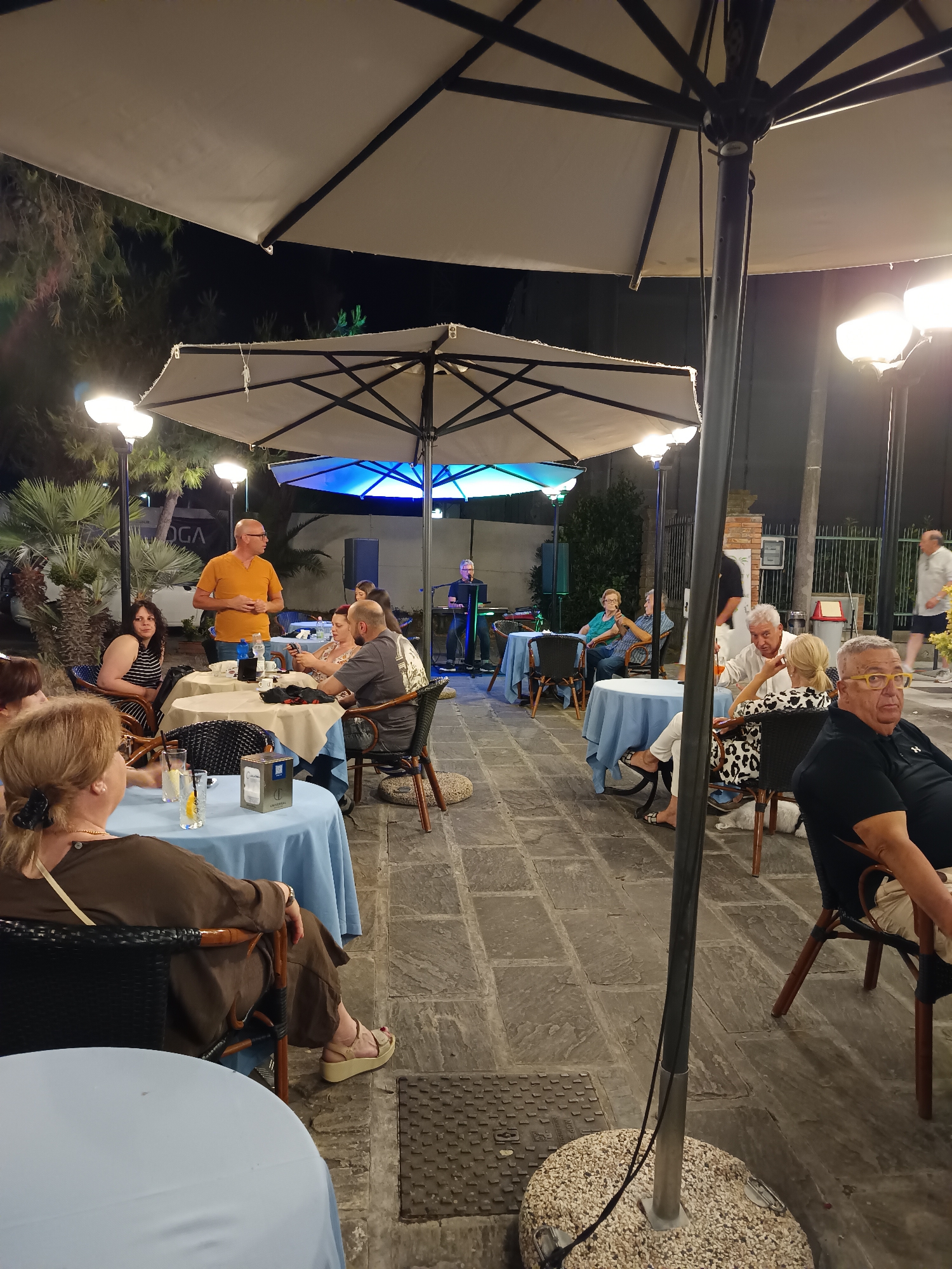 The image depicts an outdoor seating area at night, likely a restaurant or cafe, under a large umbrella. Several tables are set up, some with patrons seated, while others are empty. A man in an orange shirt stands near one of the tables, addressing a group. In the background, a musician is playing an accordion. The ground is paved with stone tiles, and there are palm trees and foliage visible. The overall atmosphere suggests a social gathering with entertainment. The scene is consistent with the outdoor dining areas found in the city of Roseto degli Abruzzi, Italy, particularly along its promenade or in its main squares during the evening.