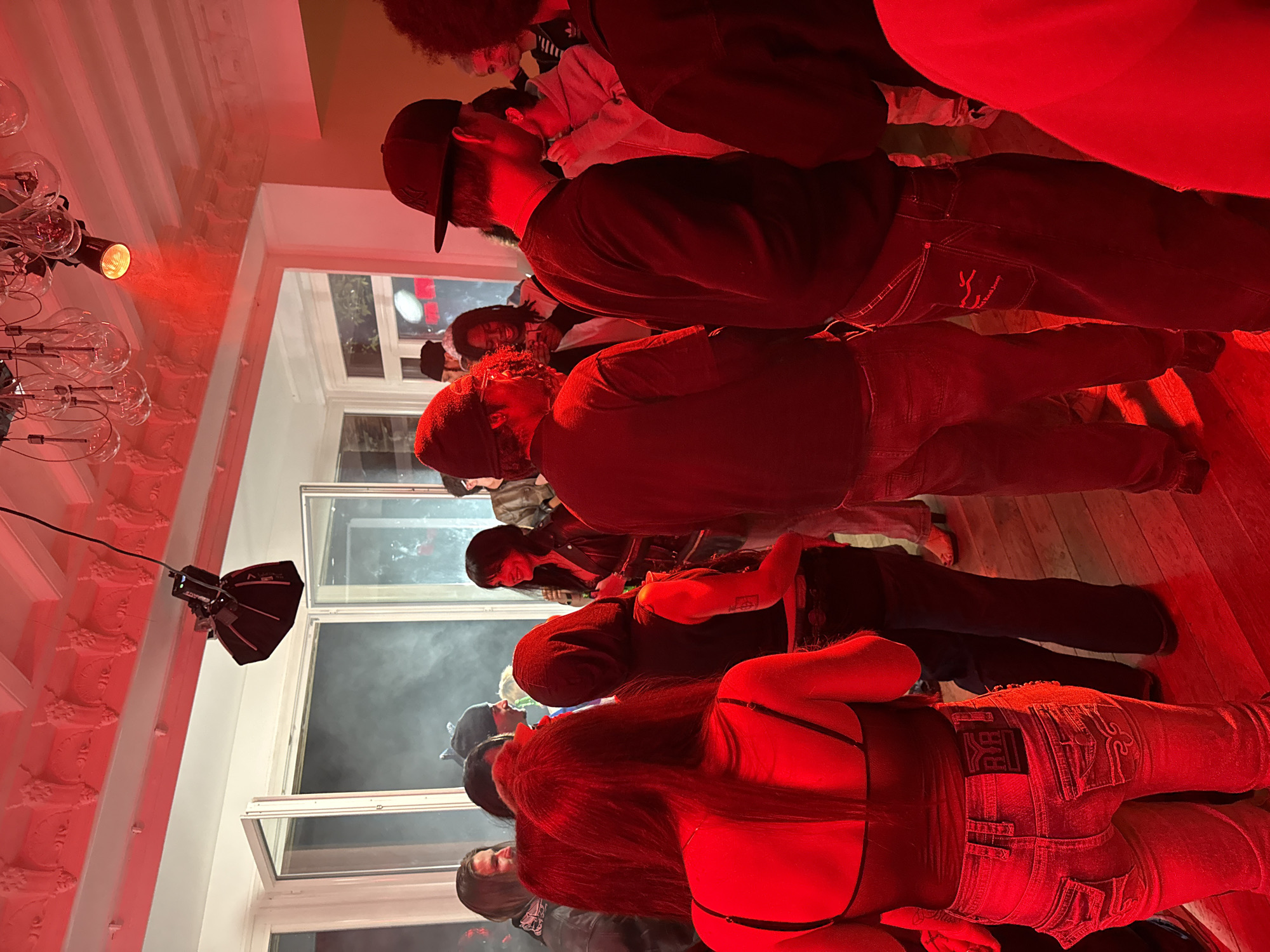 A photograph taken indoors under red lighting shows a crowd of people in what appears to be a party or gathering. The room has windows to an exterior, and a hanging light fixture with multiple bulbs is visible. An overhead stage light also illuminates the scene. The people are dressed in casual clothing. The photograph was taken in Palaiseau, France.