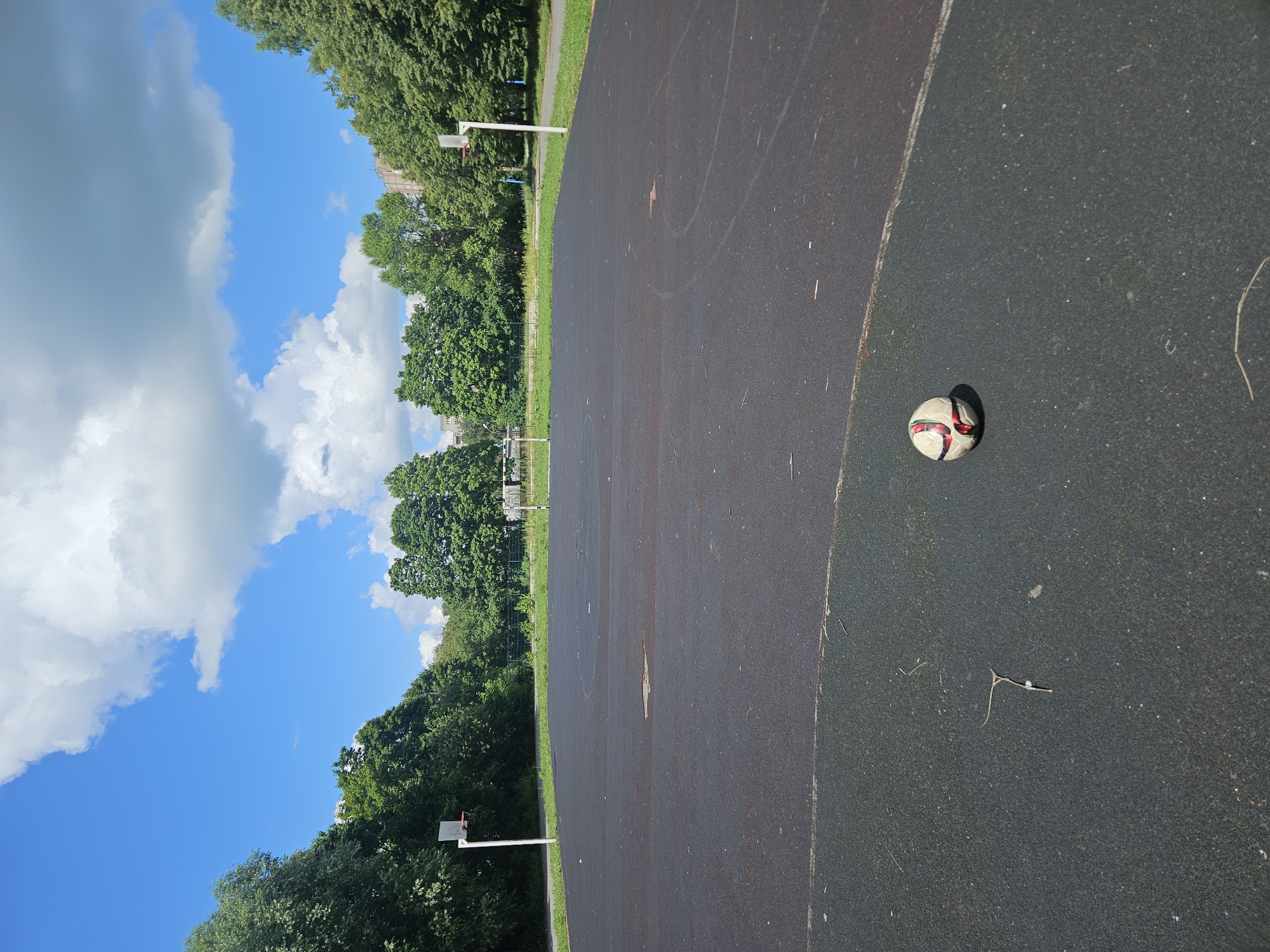 An outdoor basketball and soccer court with a worn, dark grey surface is seen under a bright blue sky with scattered white clouds. Trees line the perimeter of the court, and a few buildings are visible in the distance. Two basketball hoops are positioned at opposite ends of the court. In the foreground, a soccer ball with red and black accents rests on the court. The scene appears to be in a park or recreational area within the city of Vladimir, Russia.