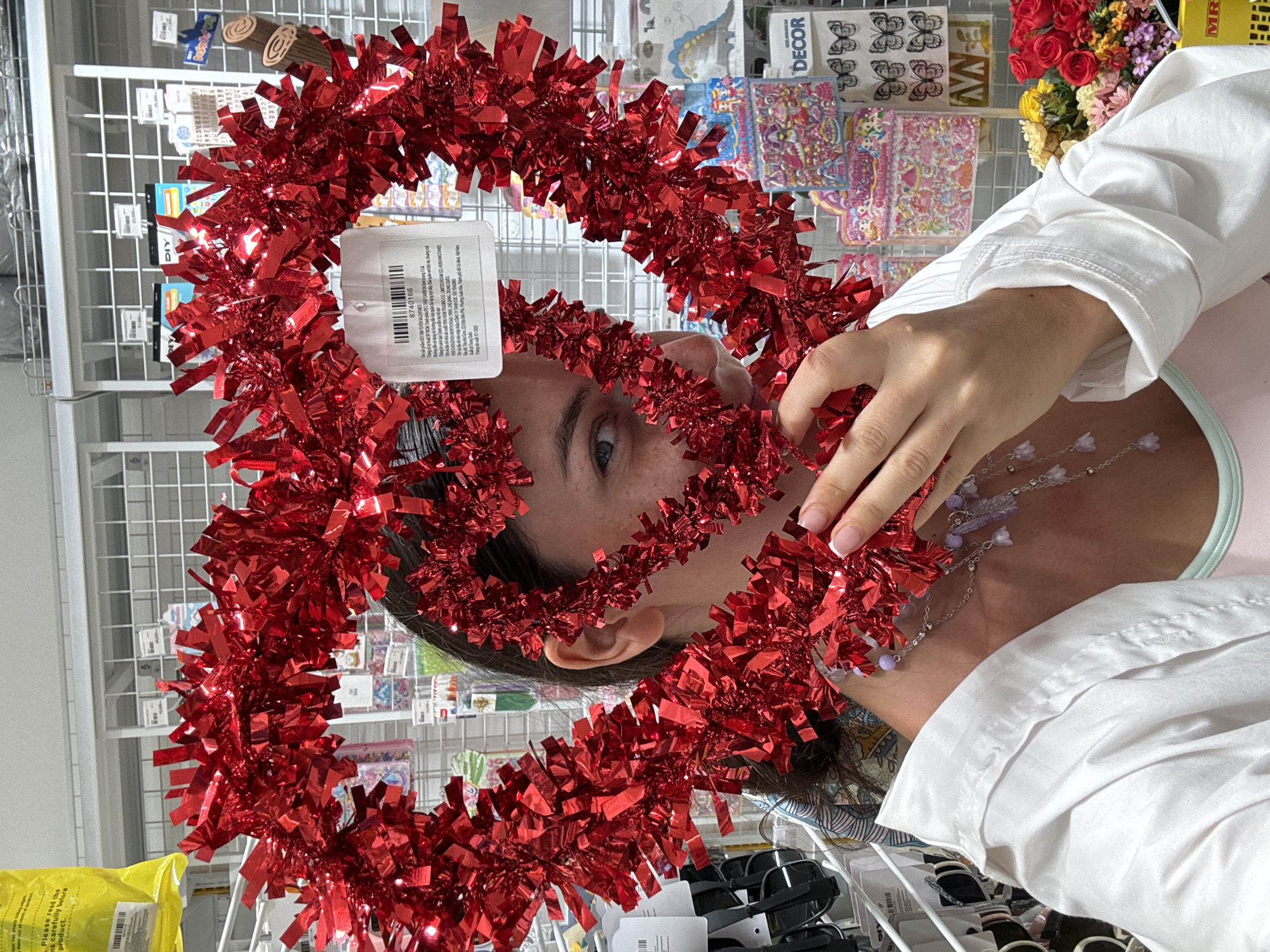 A young woman with brown hair, partially obscured by a large, red, heart-shaped tinsel decoration, is shown in a store. She is wearing a white collared shirt over a pink top and a silver necklace with purple beads and charms. Her right hand is raised, touching the tinsel. Behind her, shelves are stocked with various items, including stickers, craft supplies, and flowers in the lower right corner. The store appears to be located in Phu Thuy Ward, Vietnam.
