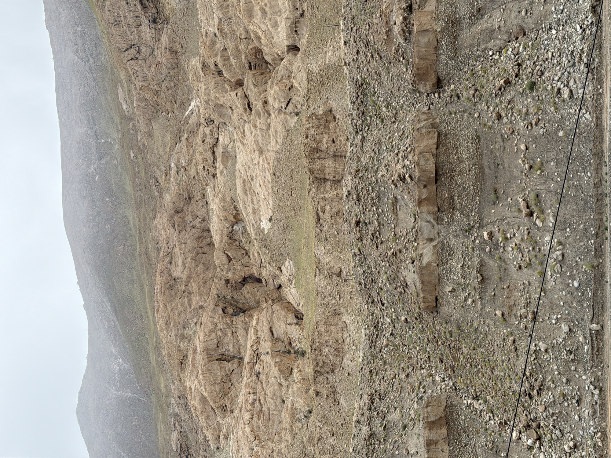 The image shows a rocky, mountainous landscape in the city of Kungyamdo, India. The terrain is characterized by steep slopes covered in loose rocks, gravel, and sparse vegetation. The dominant colors are earthy tones of brown, beige, and gray. In the foreground, a black cable runs horizontally across the frame, likely a power line or utility wire. The background features a large, imposing mountain range with a hazy, overcast sky, suggesting potentially damp or foggy weather conditions. The rocky formations exhibit signs of erosion, with visible layers and crevices. There are no people or animals visible in the image. The overall scene conveys a sense of rugged, natural wilderness.