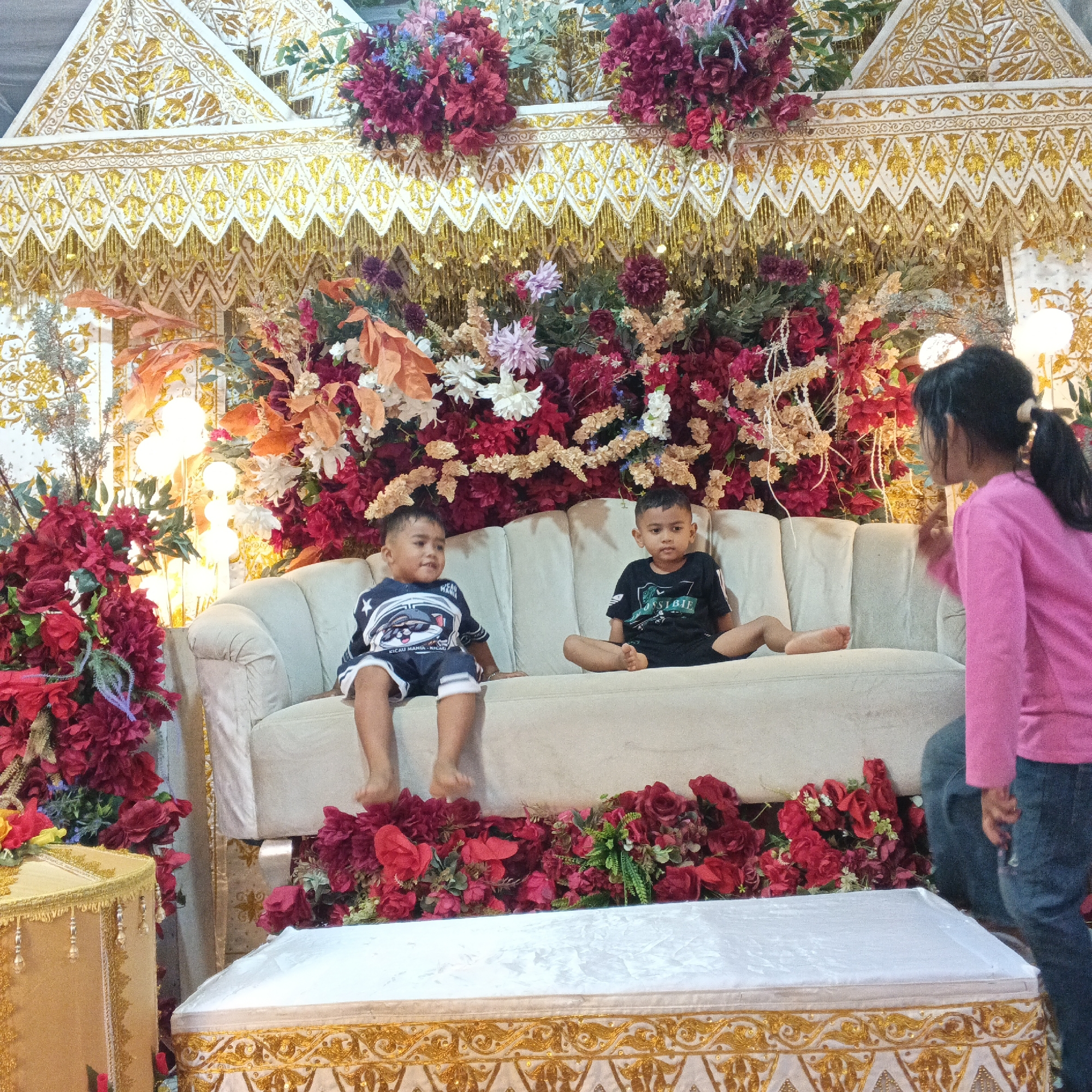 In an indoor setting, two young boys are seated on a cream-colored sofa, facing the camera. The boy on the left wears a blue and white shirt with a graphic print and blue shorts. The boy on the right wears a black t-shirt with a white graphic and appears to be sitting with his legs crossed. A young girl in a pink long-sleeved shirt and blue jeans stands to the right of the sofa, facing away from the camera and looking towards the seated children.
The backdrop is elaborately decorated with numerous red and pink artificial flowers, interspersed with gold and cream-colored fabric draping. There is a white structure with gold ornate patterns at the top, possibly part of a stage or backdrop. In the foreground, a white object with a gold patterned trim, resembling a coffin or a ceremonial box, is positioned in front of the sofa. Another gold decorative box is visible on the far left. The lighting appears to be artificial, with bright lights visible behind the floral arrangements. The overall scene suggests a festive or ceremonial occasion, possibly a wedding or cultural event. The location is identified as Unknown, Unknown.