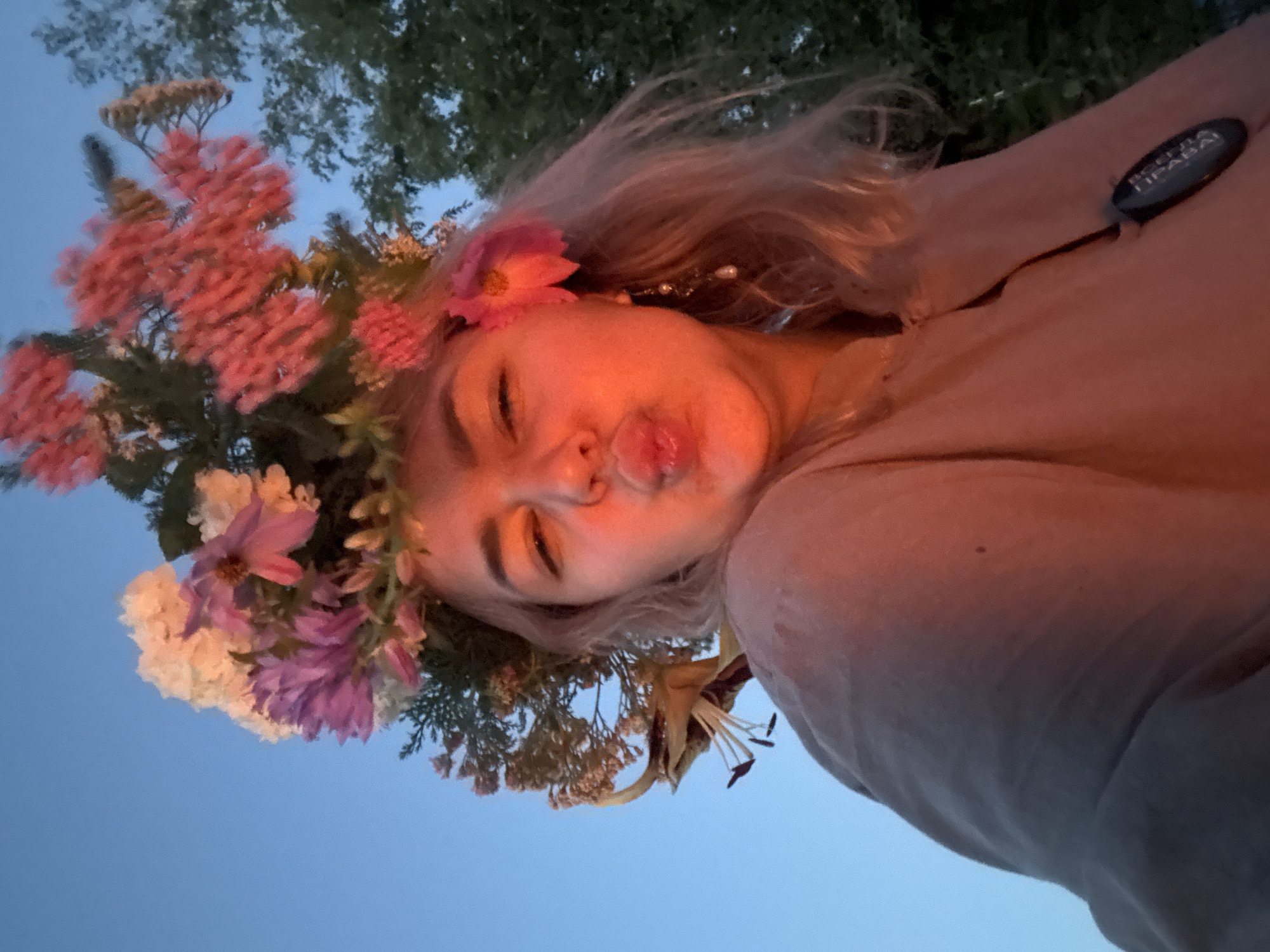 A young woman is depicted in a selfie, wearing a flower crown and a light-colored garment. She is puckering her lips, possibly for a kiss, and her right eye is partially closed. The flower crown is a prominent feature, adorned with pink, white, and purple blossoms, as well as greenery and a yellow lily. She has blonde hair and is wearing an earring on her right ear. A dark, oval-shaped object with text is attached to her garment on her left side. The background consists of a clear blue sky and trees. The lighting suggests it is either sunrise or sunset, casting a warm, orange glow on her face and the upper part of her clothing. The overall setting appears to be outdoors. The specific location in Birobidzhan, Russia, cannot be determined from the image.