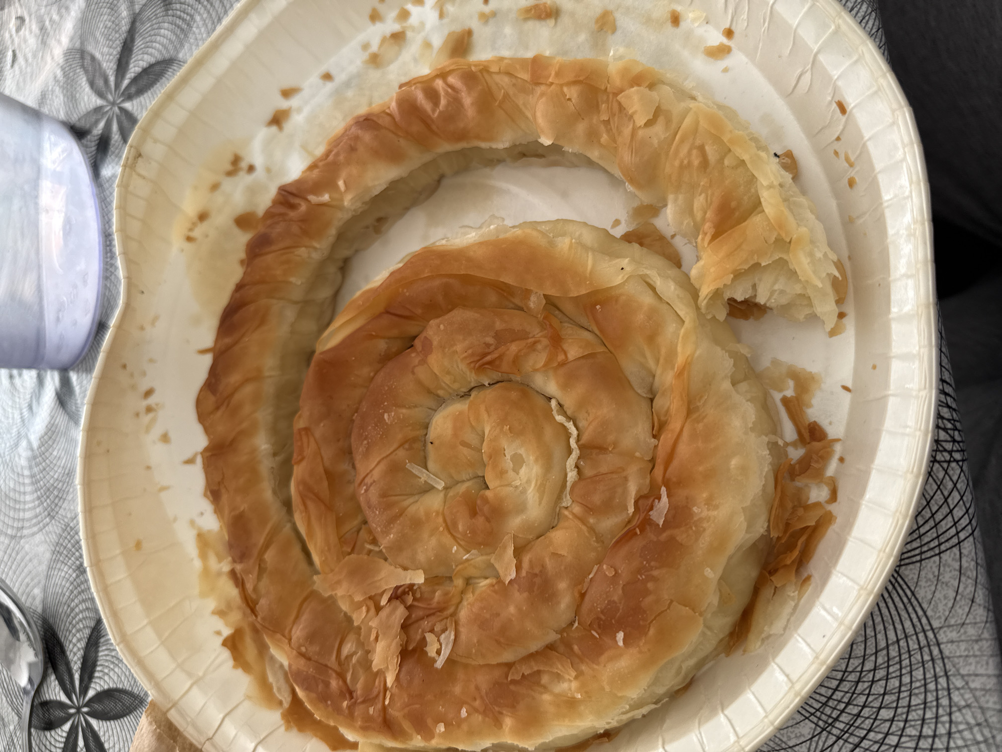 A baked spiral pastry, golden brown in color, sits on a white, fluted disposable plate. A portion of the pastry has been broken off. The plate is placed on a surface with a gray and white patterned fabric. Part of a clear plastic container is visible to the left of the plate. The location is not discernible from the image. No people, actions, or interactions are depicted.