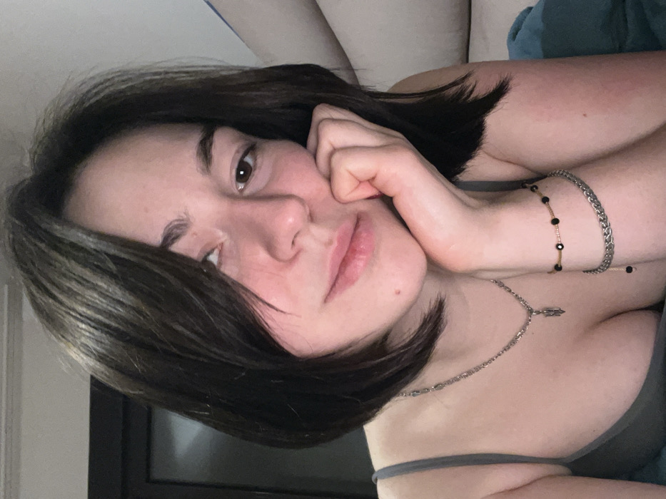 A young woman with short dark hair rests her cheek on her hand. She wears a gray tank top and a silver necklace with a small charm. Two bracelets adorn her wrist; one is a silver chain, and the other features black beads. Her expression is neutral, and she gazes towards the camera. The background includes hints of a window, a blanket, and possibly a headboard.