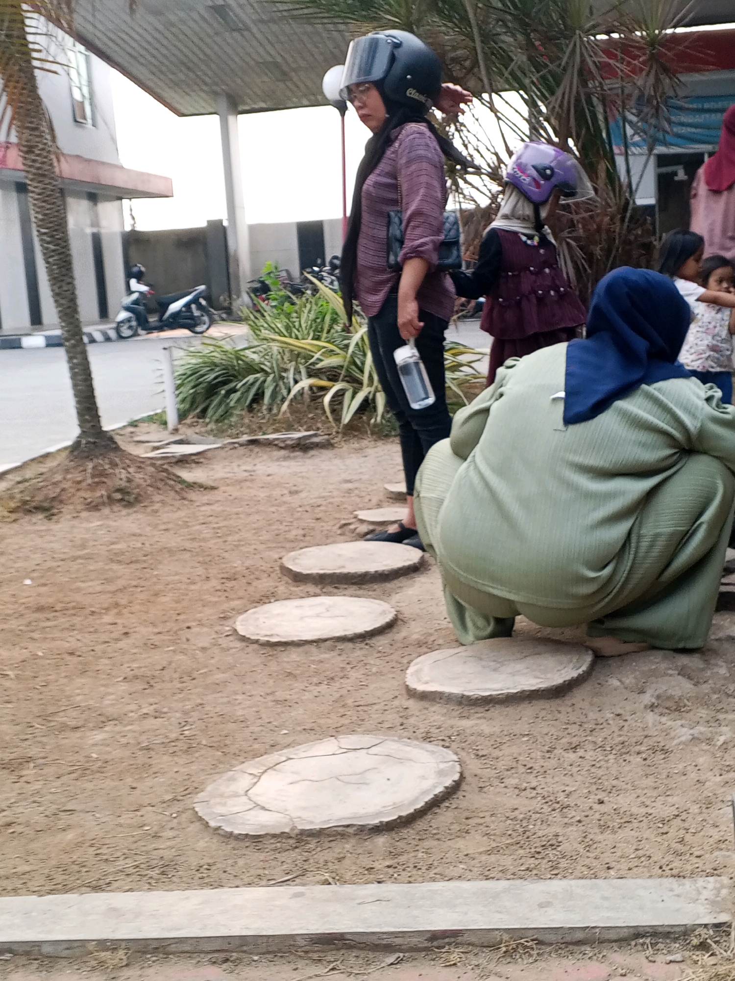 In Ciruas, Indonesia, a group of people are gathered near a building and a palm tree. A woman wearing a purple long-sleeved shirt and black pants, with a black helmet and black bag, stands holding a water bottle. To her right, a child in a purple dress and a purple helmet is visible. Further to the right, another child is partially visible wearing a white shirt with a floral pattern. In the foreground, a person in a green outfit with a blue headscarf is squatting. Several circular stepping stones are laid out on the dirt path. In the background, motorcycles are parked, and the edge of a building with a roof structure is present.