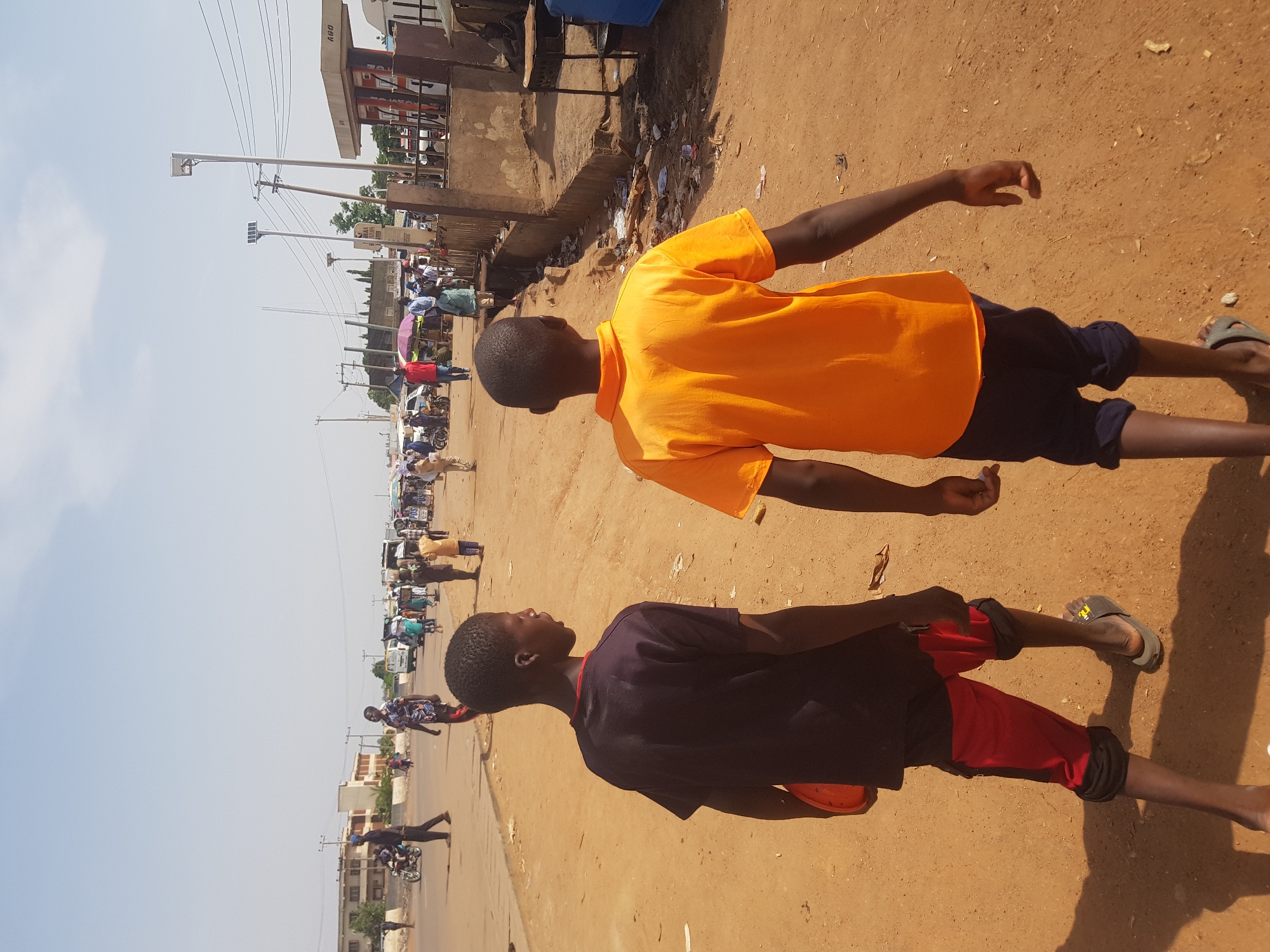 This image captures a vibrant street scene in Tukur Tukur, Nigeria, on a bright, sunny day. The primary subjects are two young males, likely boys or teenagers, positioned in the foreground on an unpaved, reddish-brown dirt road. The boy on the left wears a dark blue/black t-shirt with red shorts and sandals, while the boy on the right is in an orange t-shirt, dark blue shorts, and flip-flops. Both appear to be walking or standing on the side of the busy road.
In the mid-ground and background, the street extends into a bustling area. Numerous people are visible, many congregating around what appear to be market stalls or small businesses. Various vehicles, including motorcycles and what look like minibusses or vans, are present, indicating a lively commercial or residential thoroughfare. Utility poles with multiple power lines run along the street, and modern streetlights, some equipped with visible solar panels, are also installed.
On the right side of the road, buildings line the street, including a structure that appears to be a petrol station, with the text "R&O" discernible on its sign. The sky is clear and light blue, and strong shadows cast by the subjects and objects suggest bright daylight, likely mid-morning or early afternoon. The overall scene portrays everyday life and commerce in a West African town.