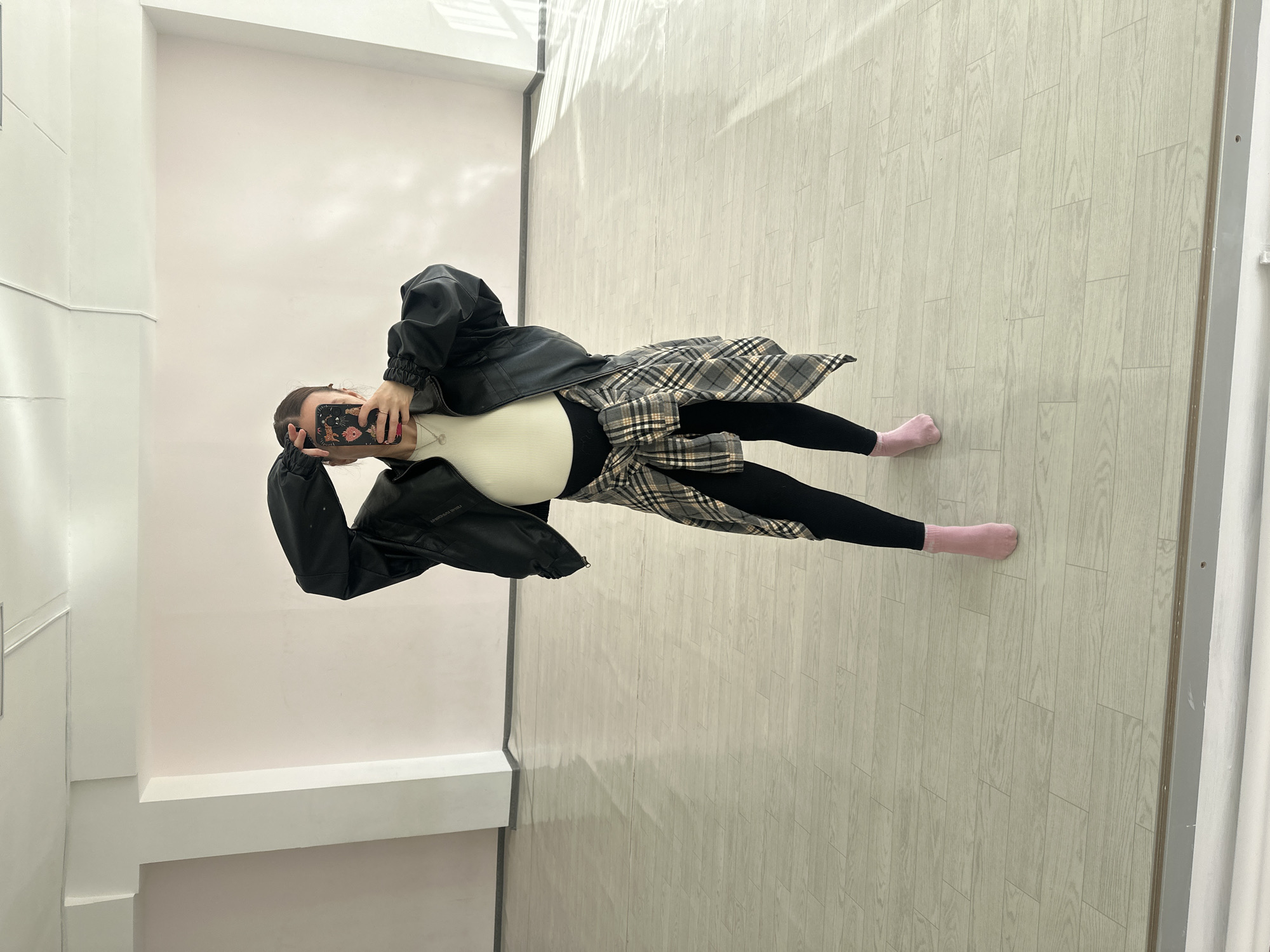 A woman stands indoors against a white wall, holding a phone in her left hand, obscuring her face. She wears a white top, black leggings, and pink socks. A black leather jacket and a plaid shirt are draped around her torso. The floor is covered in light-colored wood-patterned flooring. The location is unknown and cannot be identified as Votkinsk, Russia based solely on this image.