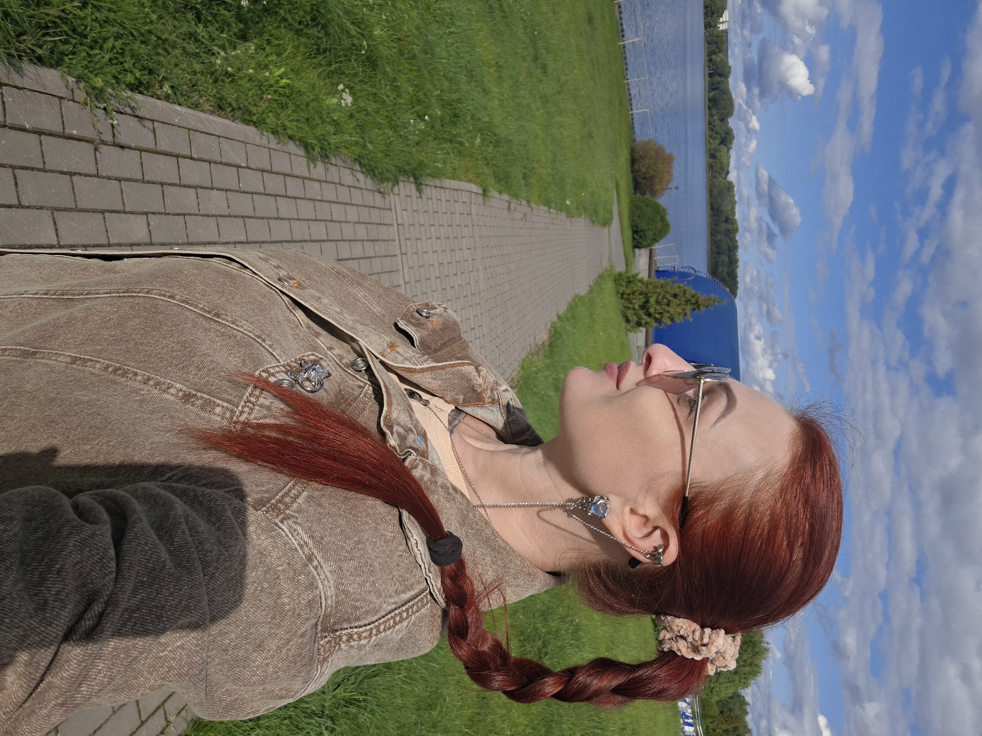 A medium shot captures a woman with auburn hair styled in a braid, wearing sunglasses and a denim jacket. She is standing outdoors on a paved pathway next to a grassy area. In the background, a body of water, trees, and a large blue structure are visible under a partly cloudy sky. The scene appears to be a park or recreational area, possibly near a lake, in the city of Myadzyel, Belarus.