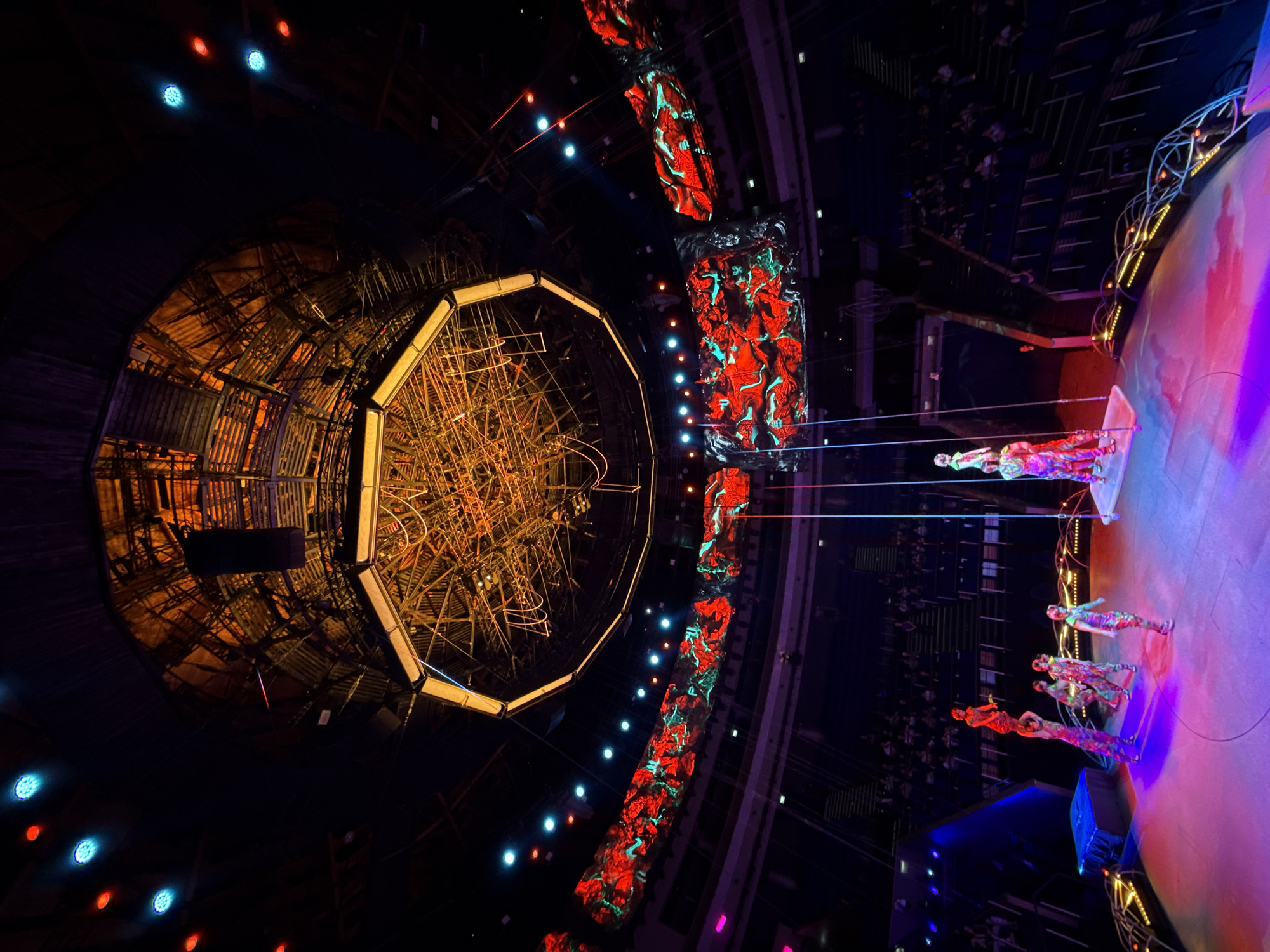 The image shows a performance at the Karen Demirchyan Sports and Concerts Complex in Yerevan, Armenia. Performers in brightly colored costumes are on a circular stage lit with pink and blue hues. A large, ornate, geometric structure hangs above the stage, illuminated in yellow and orange tones. Red and green patterned screens flank the stage, and lights are arrayed around the performance area. Audience members are visible in the background.