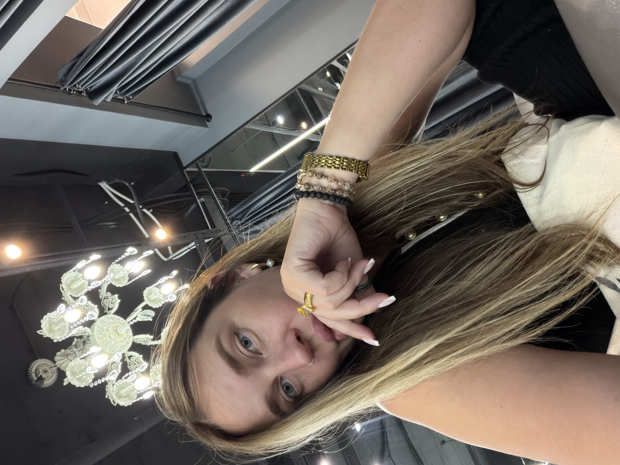A person is positioned with their face directed towards the camera, their left hand raised to their mouth, and fingers touching their lips. The person has fair skin, light blue eyes, and light brown hair with blonde highlights. On their left wrist, they are wearing a gold watch and several beaded bracelets. They are wearing a gold ring with a yellow stone on the ring finger of their left hand. They also have a stud earring in their left ear and a nose piercing.
The person is wearing a black top with white trim. Behind the person and to their left, there is an ornate chandelier with multiple light bulbs. In the background, there are gray curtains hanging from a track system. The location is Moscow, Russia.