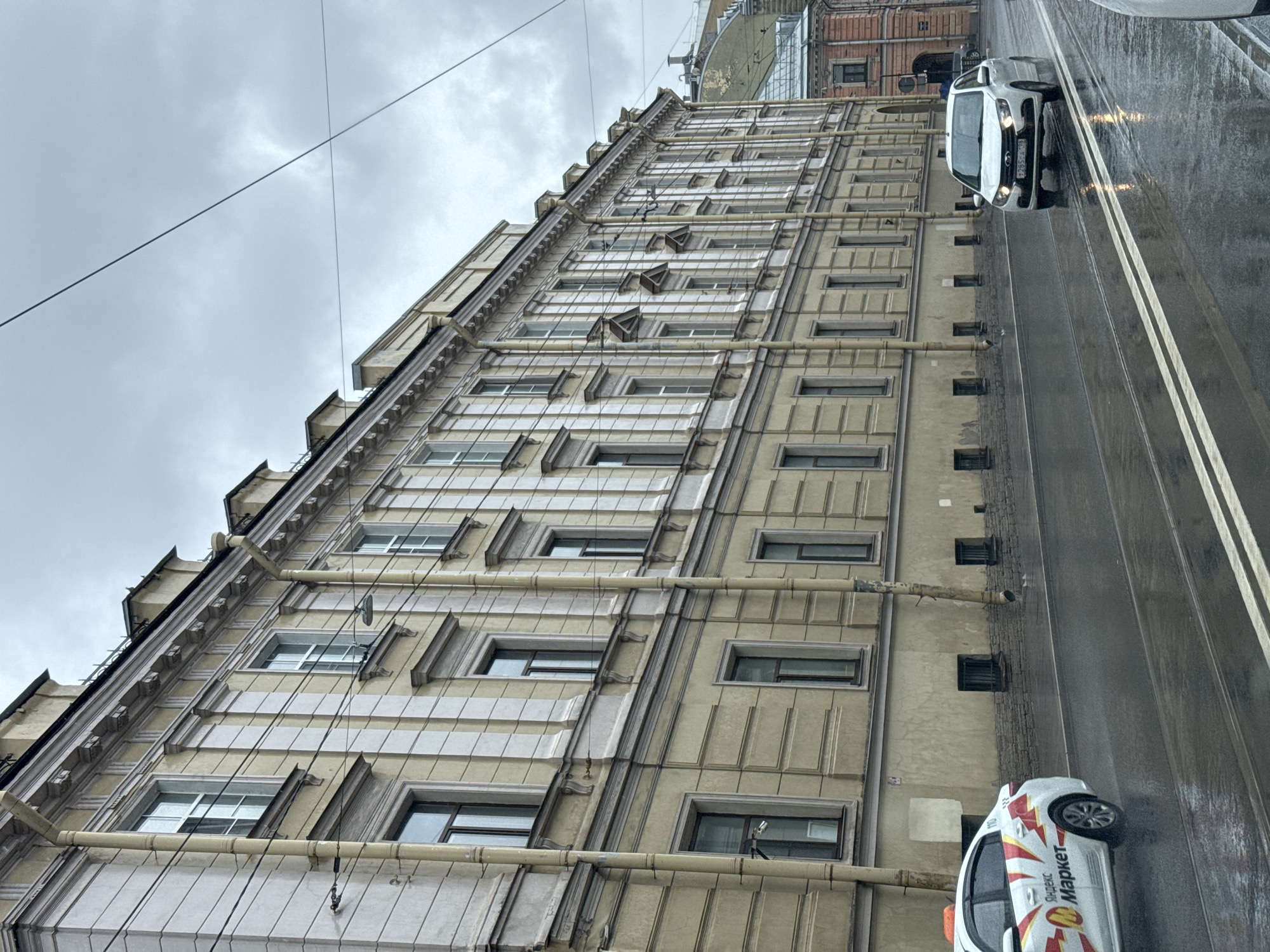 The image depicts a cloudy day on a city street in Saint Petersburg, Russia, likely on a wide avenue given the width of the road and the presence of tram tracks. The street is wet, indicating recent or ongoing rain. A long, multi-story building with a pale yellow facade and numerous windows lines the left side of the street. The building features classical architectural details, including pilasters, ornate window frames, and a decorative cornice. Vertical drainpipes run along the facade.
In the foreground on the left, a white car with red and yellow branding, identifying it as a "Яндекс.Маркет" (Yandex.Market) taxi, is driving away from the viewer. On the right side of the street, a white sedan is approaching, its headlights on. The road has double white lines indicating lanes. In the background, other buildings with differing architectural styles are visible, including one with a reddish-pink facade. Overhead wires are strung across the scene. The sky is overcast and gray, consistent with typical Saint Petersburg weather. There are no visible pedestrians. The overall impression is a subdued, everyday urban scene on a wet day.