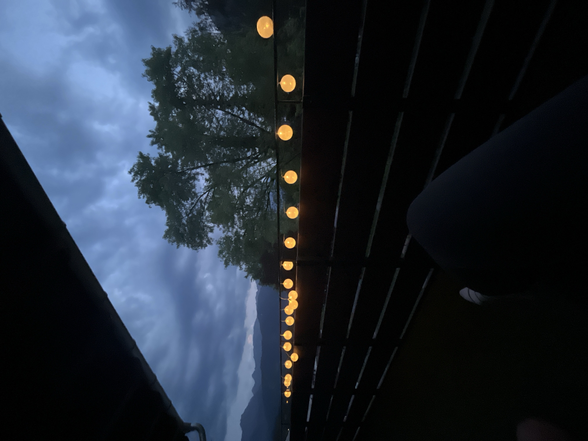 The image depicts an outdoor scene at dusk or night, likely on a balcony or porch overlooking a mountainous landscape. A string of round, illuminated fairy lights stretches horizontally across the frame, suspended above a dark wooden fence. The lights cast a warm, orange glow against the darkening sky, which is filled with dramatic, heavy clouds. In the background, silhouetted trees are visible, and beyond them, the faint outlines of mountains suggest a location in or near Garmisch-Partenkirchen, Germany. The foreground is dominated by the dark interior of the balcony, with a person's legs and feet, clad in dark trousers and white socks, visible in the lower right corner. The overall atmosphere is moody and serene, hinting at a quiet evening outdoors.