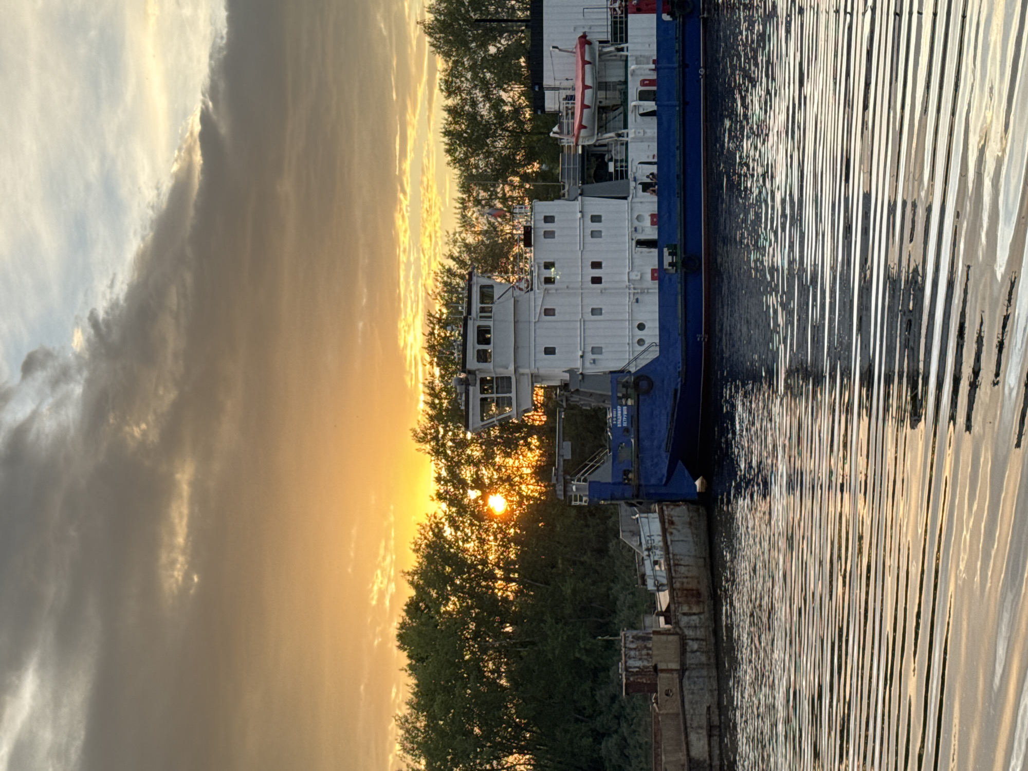 The image depicts a serene sunset over the Kama River in Chaikovsky, Russia. The sky is a canvas of warm oranges and yellows, with dramatic clouds tinged with gray. The sun, a bright orb, peeks through a dense line of green trees on the riverbank, casting a golden glow on the water. Moored along the bank are two barges. The larger, closer barge is predominantly blue with a white superstructure. It features multiple decks, windows, and a life raft. The smaller, more weathered barge is to its left. The rippling surface of the Kama River reflects the warm hues of the sunset and the dark silhouettes of the barges, creating a tranquil and picturesque scene.