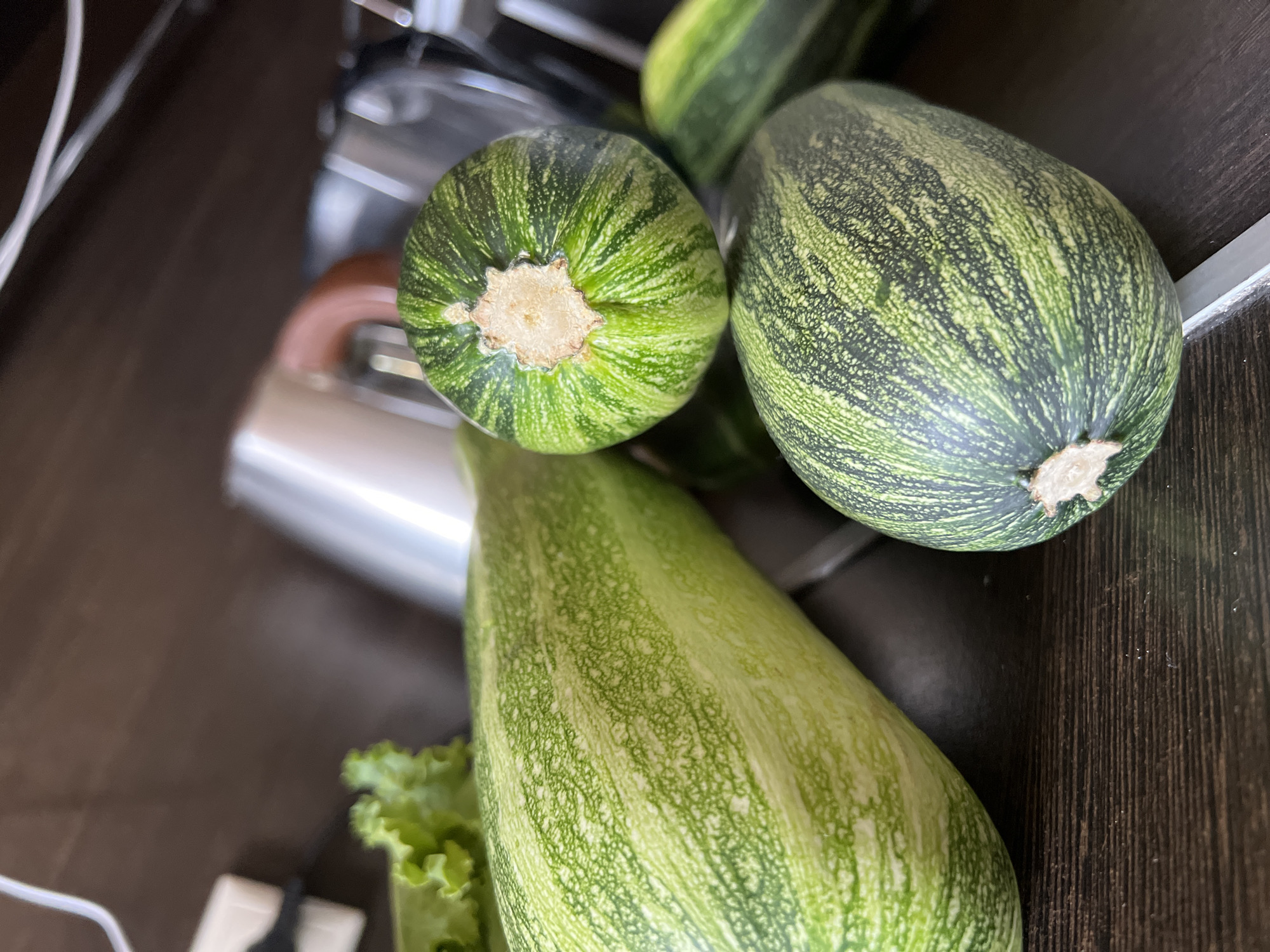 This is a close-up, slightly angled, eye-level shot of several green zucchini on a dark brown wooden surface. The zucchini exhibit varying patterns of light and dark green stripes and speckles. One large zucchini runs diagonally from the left foreground to the center. Two rounder zucchini are positioned to the right of the large one, with one slightly behind and above the other. A leaf of green lettuce is visible in the upper left corner, out of focus. In the background, out of focus, are parts of silver kitchen appliances with brown handles and white electrical cords. The scene is indoors, likely in a kitchen. There is no indication of the location being in Tolyatti, Russia.
