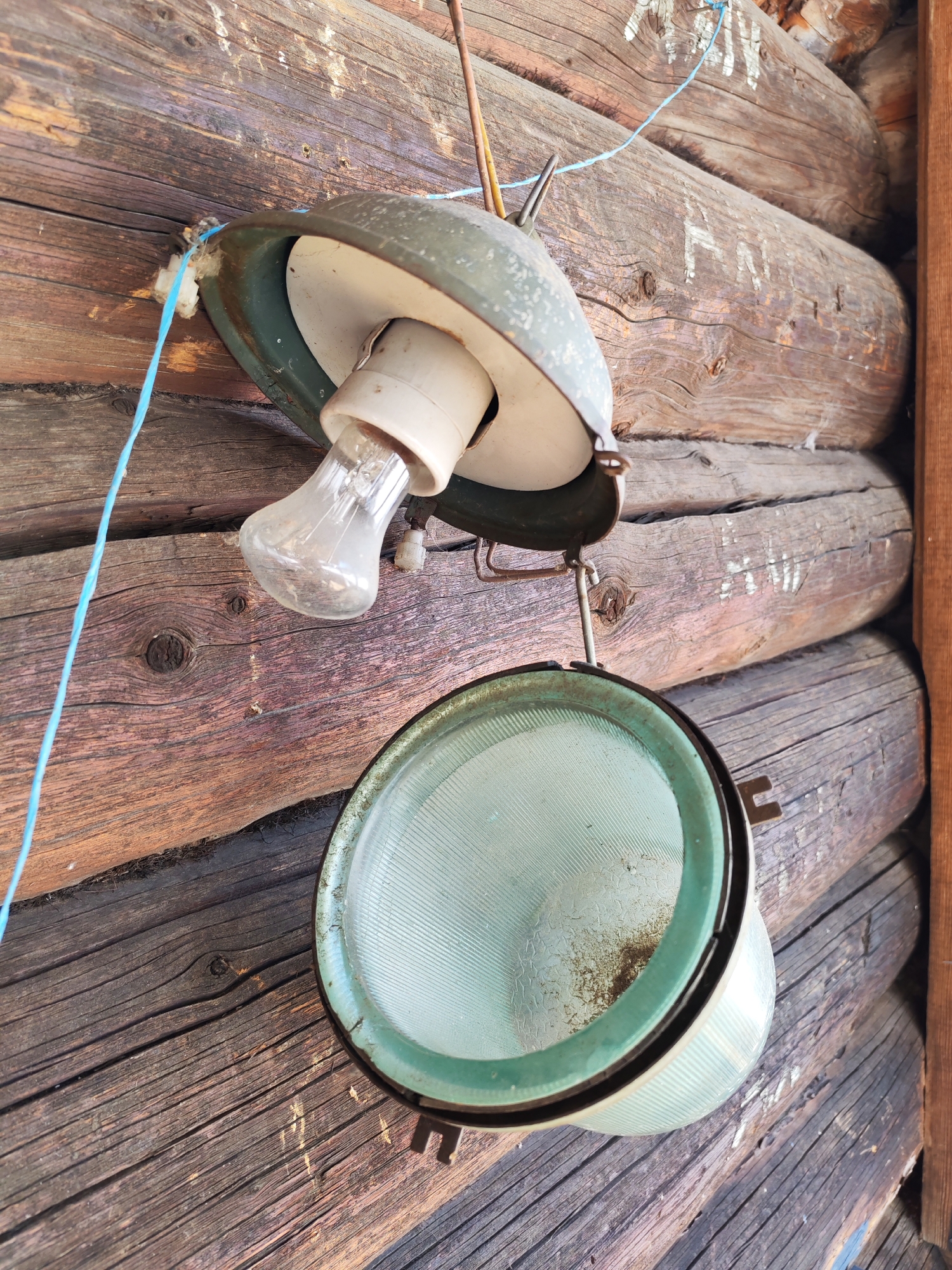 A close-up shot shows an old, potentially broken, outdoor light fixture hanging against a rough, unfinished wooden wall. The wall appears to be part of a log cabin or similar rustic structure. The light fixture consists of two main parts: an upper, conical shade, and a lower, bowl-shaped diffuser. The upper shade is olive green on the outside and white on the inside. It has a standard bulb socket holding an old-fashioned incandescent bulb. The lower diffuser is a pale blue or green color and appears to be textured. The entire fixture has a somewhat dilapidated look, suggesting it has been exposed to the elements for a considerable time. A blue cord is seen dangling near the fixture. The location is Chaikovsky, Russia.