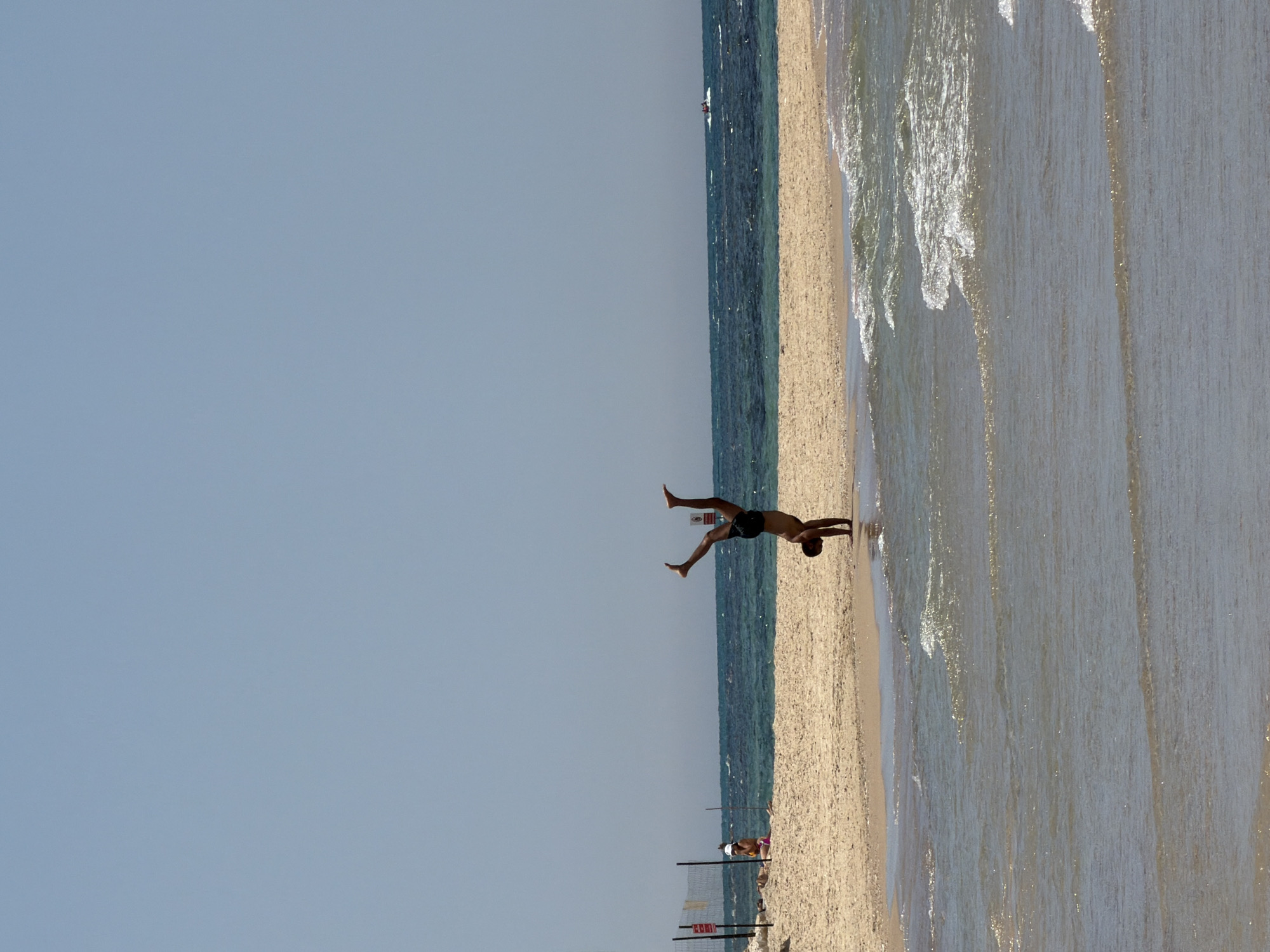 This image captures a vibrant beach scene in Tel-Aviv, Israel, under a clear, bright sky. The main subject is a fit individual, likely a man, performing an athletic pose resembling a handstand or acrobatic balance on the wet sand at the water's edge. He is wearing dark shorts and is positioned with his hands on the sand, legs spread wide in a dynamic, inverted stance.
The scene features the sparkling blue-green waters of the sea to the left, with gentle waves breaking onto the shore. The sandy beach stretches along the right side of the frame, showing both wet and dry sand textures. In the mid-ground, further down the beach, another person can be seen sitting near a low, white and black fence or netting structure, possibly marking a section of the beach.
The time of day appears to be sunny daytime, with ample light illuminating the scene, indicating fair weather conditions. The overall atmosphere is lively and active, showcasing leisure and physical activity on the beach. No legible text is visible in the image.