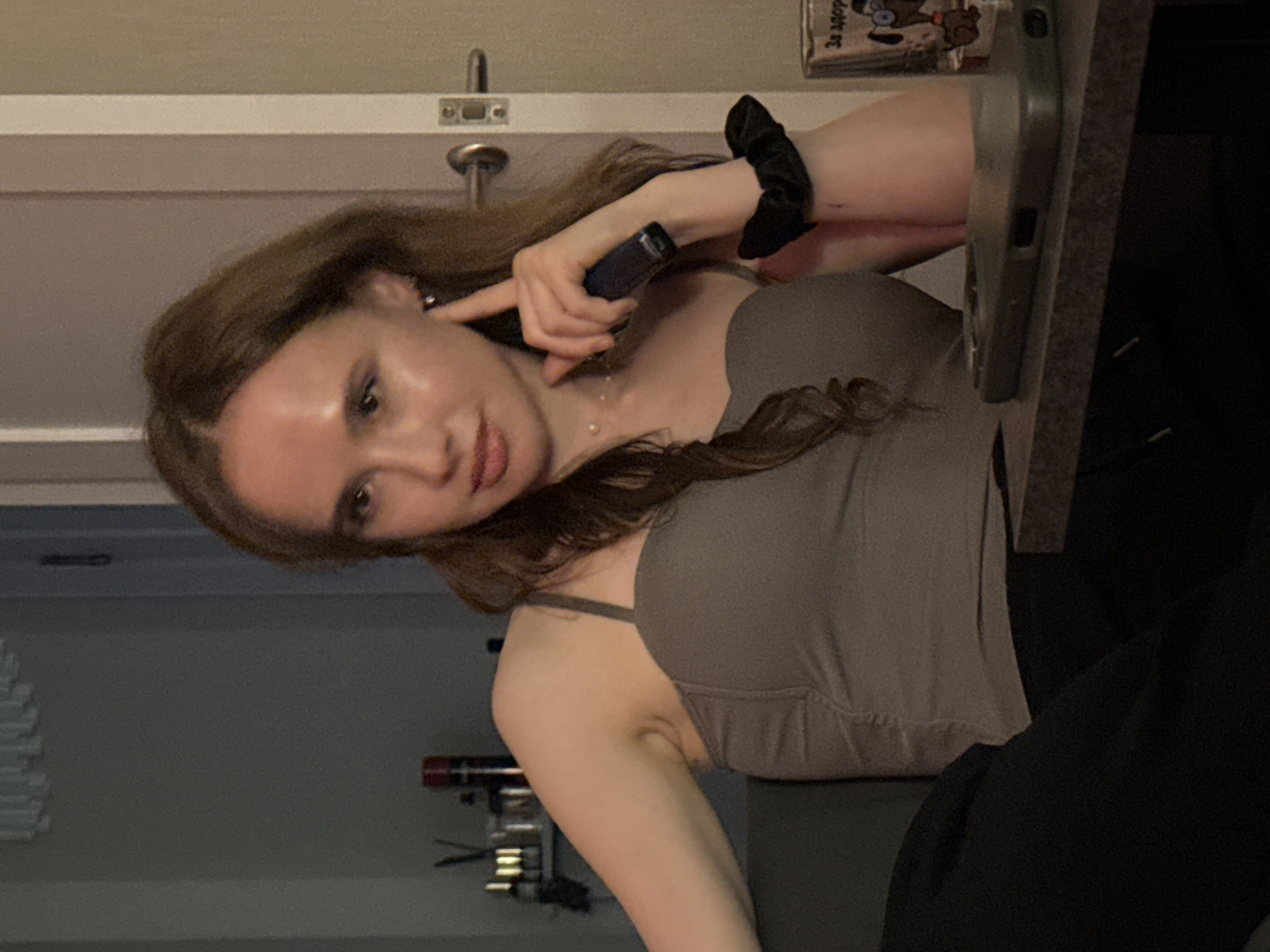 A young woman with long, light brown hair is shown in a candid, slightly angled shot, possibly taken indoors. She is wearing a grey tank top with thin straps and a black scrunchie on her wrist. Her right hand is raised, with her index finger touching her ear. She holds a small, dark cylindrical object, possibly a vape pen or a small electronic device, between her thumb and forefinger, also near her ear. Her expression is somewhat playful or curious.
In the background, a white door with a metal latch is visible. To the left, a shelf or a rack with multiple levels and objects can be seen, some of which appear to be metallic and cylindrical. A dark grey surface, possibly a desk or table, is in the foreground on the right, with a dark object resting on it. Above the woman's head, a small decorative item with cartoonish figures and text in Cyrillic is attached to the wall. The overall lighting suggests an indoor setting, possibly in the evening or with artificial light. The location context is Perm, Russia.