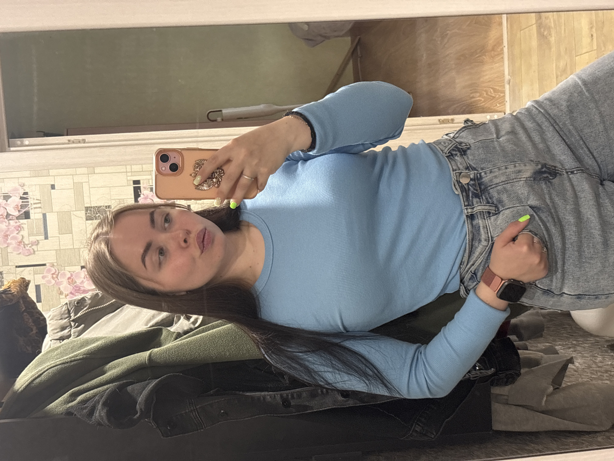 A young woman is taking a selfie in a mirror. She is wearing a light blue long-sleeved shirt and light-wash denim jeans. She has long brown hair and is holding a pink phone in her left hand. She is wearing a watch on her right wrist. The background includes a floral-patterned wall, a stack of jackets, and part of a room with a wooden floor. The room appears to be in a residential setting. There is no indication of the city of Izhevsk, Russia.
