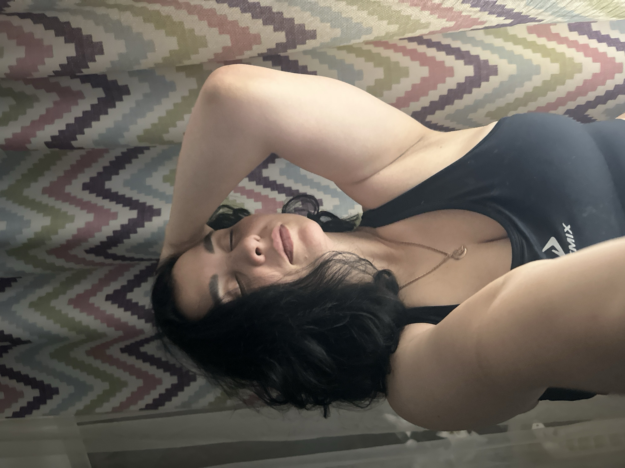 A woman is pictured from the chest up, with her eyes closed and her head tilted back, her right arm raised behind her head. She has dark, wavy hair and is wearing a dark sports bra with a silver pendant necklace. A patterned curtain is visible in the background. The image appears to have been taken indoors, possibly in a bedroom or living area.