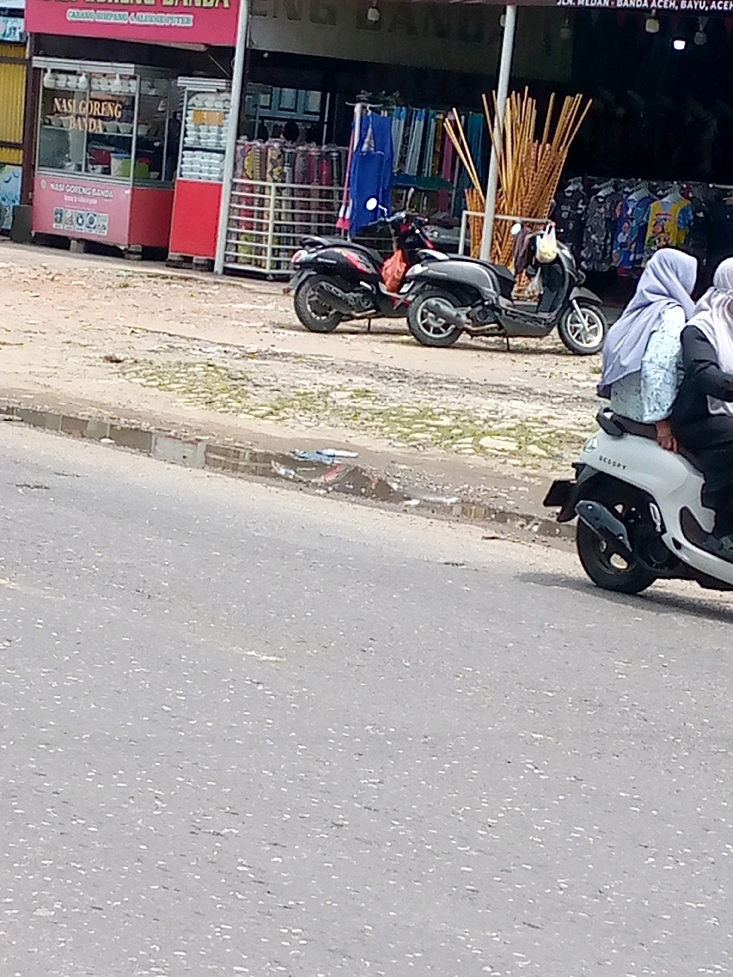 The image depicts a street scene in Keudebayu, Indonesia, with a focus on a small food stall and the surrounding environment. In the foreground, a gray asphalt road with some reflective particles is visible. To the left, a pink and red food stall with the text "NASI GORENG BANDA" is prominent, displaying various food items inside. Adjacent to the stall, several motorcycles are parked. One black scooter is in the middle ground, and a white scooter with two passengers wearing hijabs is on the right. In the background, shelves stocked with goods and an array of bamboo poles are visible. The overall scene suggests a bustling roadside area with commercial activity.