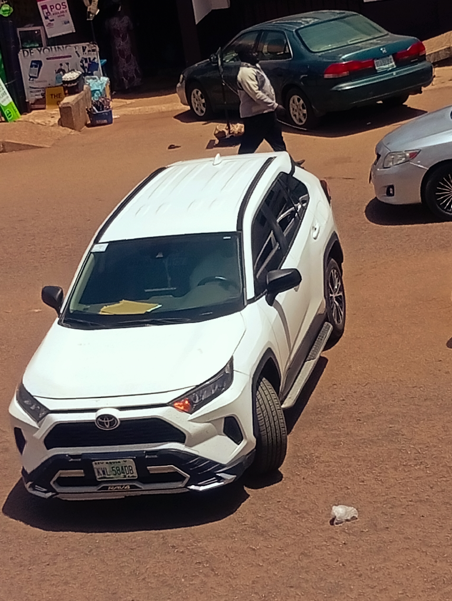 In Bukuru, Nigeria, a white Toyota Rav4 is parked on a dirt road. The car's license plate reads KWL-5840B. To the left of the Rav4, a portion of a silver car is visible. In the background, a dark green sedan is parked, and a person wearing a light-colored jacket and dark pants is walking away from it. Adjacent to the green car, a building with signage is partially visible, with one sign reading "DE YOUNG". A blue container holding various items sits on the ground near the building. The ground is covered in reddish-brown dirt, with scattered debris, including a crumpled white plastic bag near the Rav4. The lighting suggests it is daytime.