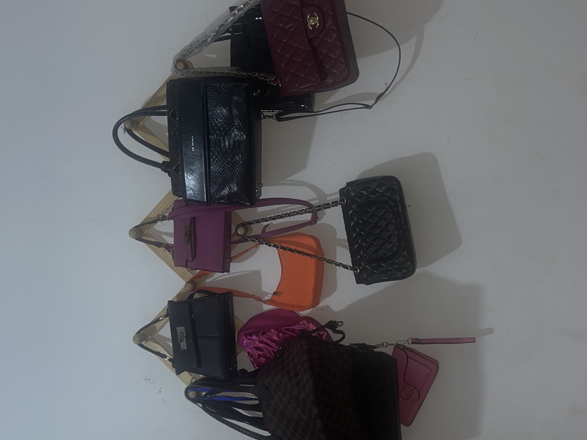 A wall-mounted wooden rack displays an assortment of handbags. The bags vary in color and style, including black, purple, orange, pink, and burgundy. Some of the bags are quilted, while others have smooth textures. The scene is indoors against a plain white wall. The location is not specified as being in Abraka, Nigeria.