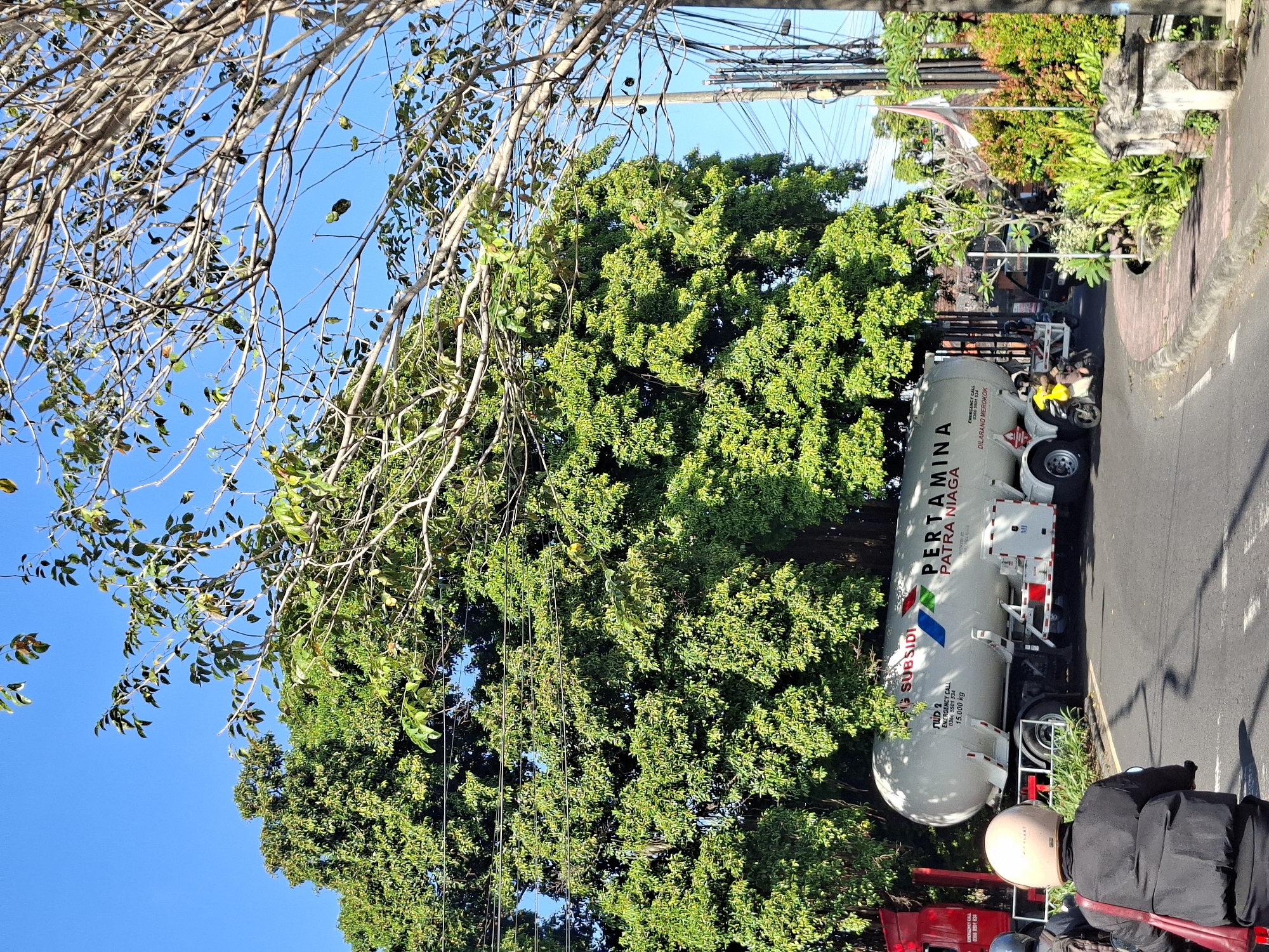 In Banjarangkan, Indonesia, on a clear, sunny day, a large white Pertamina Patra Niaga tanker truck is parked on a paved road, partially obscured by a dense green tree. The truck's tank is marked with the Pertamina logo and text, including "SUBSIDI," "EMERGENCY CALL," and "DILARANG MEROKOK" (meaning "No Smoking"). A red truck is partially visible to the left of the tanker. In the foreground, two people on motorcycles are riding away from the camera. The person in front is wearing a black jacket and a light-colored helmet, with a red bag slung over their shoulder. The person behind them is wearing a grey helmet. The road curves to the right, with a sidewalk and lush vegetation bordering it. Power lines and poles are visible in the background, along with a national flag and more trees.