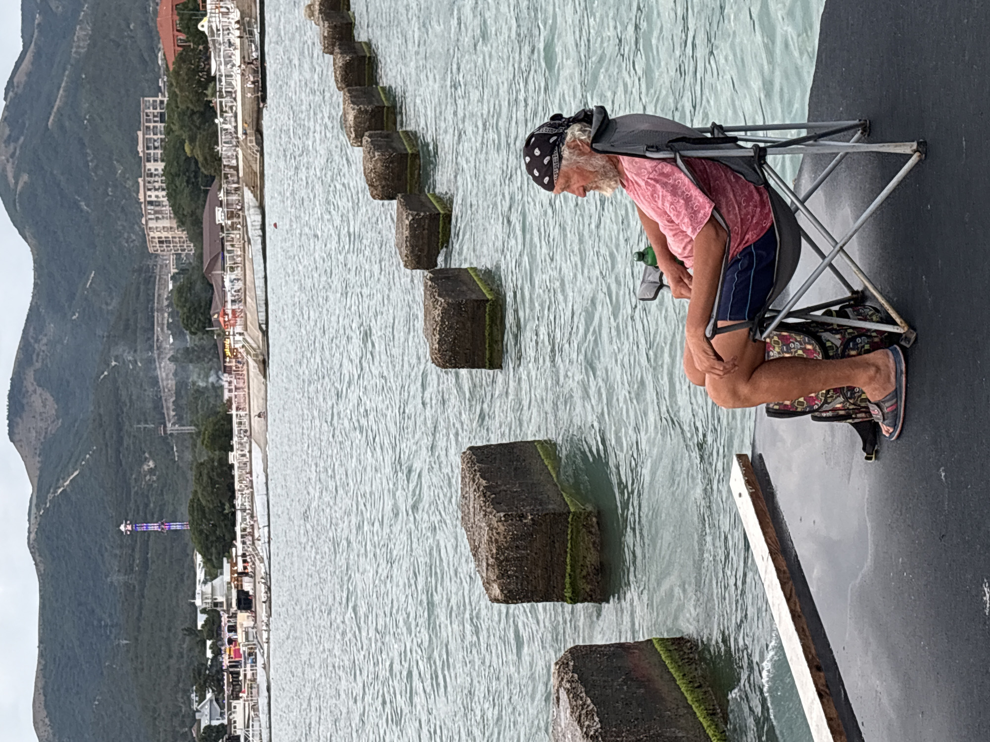 A Caucasian man with a beard and wearing a bandana sits in a folding chair on a pier in Novorossiysk, Russia. He is wearing a pink t-shirt and blue shorts. Beside him is a backpack and a green bottle. The pier is black, and there is a wooden plank in the foreground. The sea is calm and blue, with a line of concrete blocks in the water extending into the distance. In the background, there is a coastal town with buildings and a tall tower, backed by green hills under a cloudy sky.