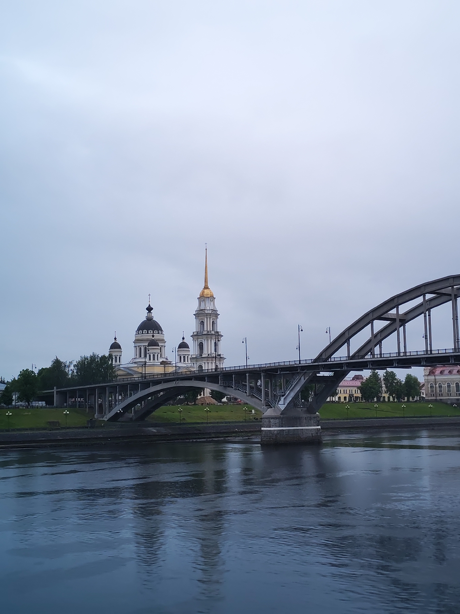 A bridge spans a body of water in Bol'shie Mkhi, Russia. The bridge is made of concrete and metal. A church with black domes and a gold steeple is visible on the left side of the bridge. The sky is overcast.
