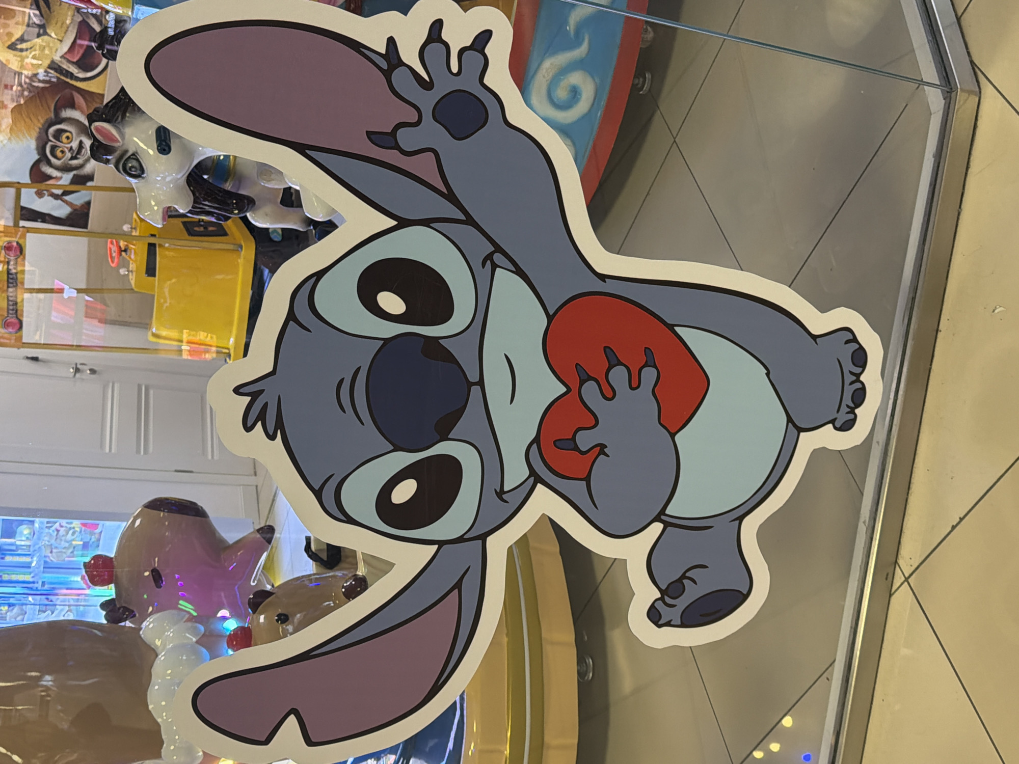 The image shows a large cutout of the cartoon character Stitch holding a red heart. Stitch is blue with large ears and big eyes. He is standing on one leg with his arms outstretched. The cutout is placed in front of a glass window, revealing a store interior with various arcade-style rides and decorations in the background. These include colorful carousel animals, a yellow ride with a steering wheel, and possibly other cartoon characters visible in the distance. The floor appears to be tiled with a light-colored material. The scene suggests a playful and entertainment-oriented environment, likely a children's play area or arcade. The mention of Vologda, Russia, is not supported by visual evidence in the image itself.