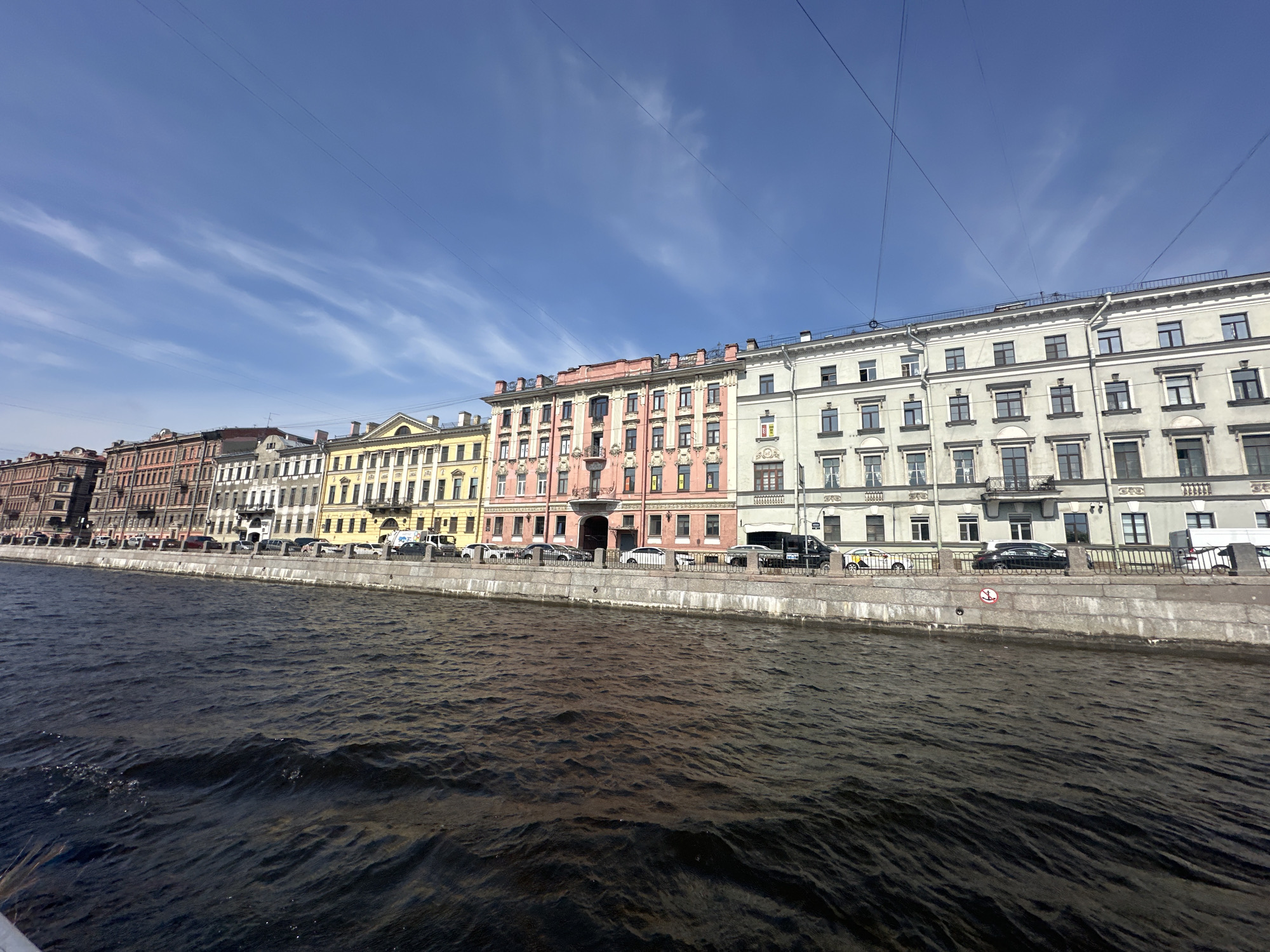 The image depicts a waterfront view of historical buildings in Saint Petersburg, Russia. The architecture showcases a variety of styles and colors, ranging from brown and yellow to pink and gray, with ornate facades and multiple windows. A concrete embankment borders the water, separating it from the buildings and the street where cars are parked. Overhead wires stretch across the sky, contrasting with the clear blue sky marked with wispy clouds. The water is dark and rippled, suggesting a moving current.