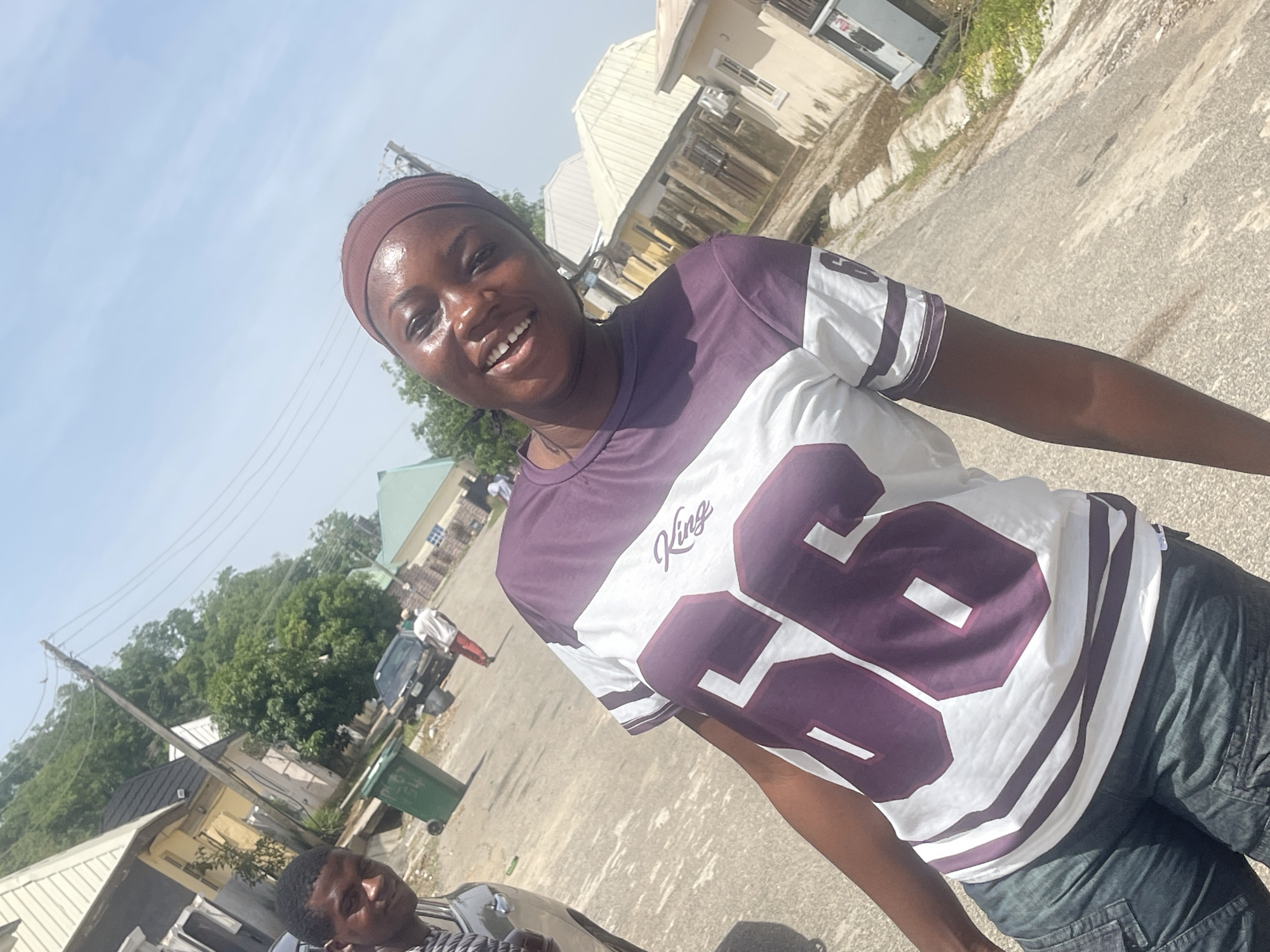 A medium shot captures a woman with a brown headband and a wide smile, posing outdoors. She wears a shirt with a large number "86" design on it and dark pants. The background reveals a street in Abuja, Nigeria, with residential buildings, vehicles, trees, and a person walking. A man's head and shoulder are also visible in the lower left corner of the image.
