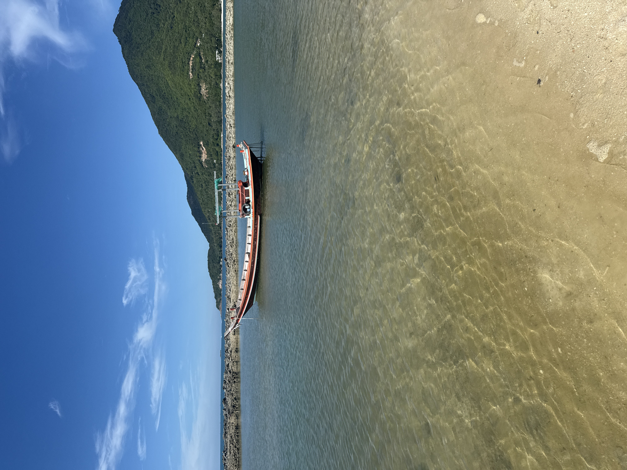 A longtail boat is moored in clear, shallow water near a rocky shoreline. The boat is traditional in design with a pointed bow and stern, and a wooden canopy covering the central part of the vessel. It has a red and white color scheme along its hull. The water is transparent, allowing the sandy bottom and small stones to be visible. In the background, a large, tree-covered mountain rises under a clear blue sky with a few wispy clouds. The scene appears to be a tranquil coastal area within ตำบลเกาะพะงัน, Thailand. The image is captured from a low angle, looking towards the water and the boat.