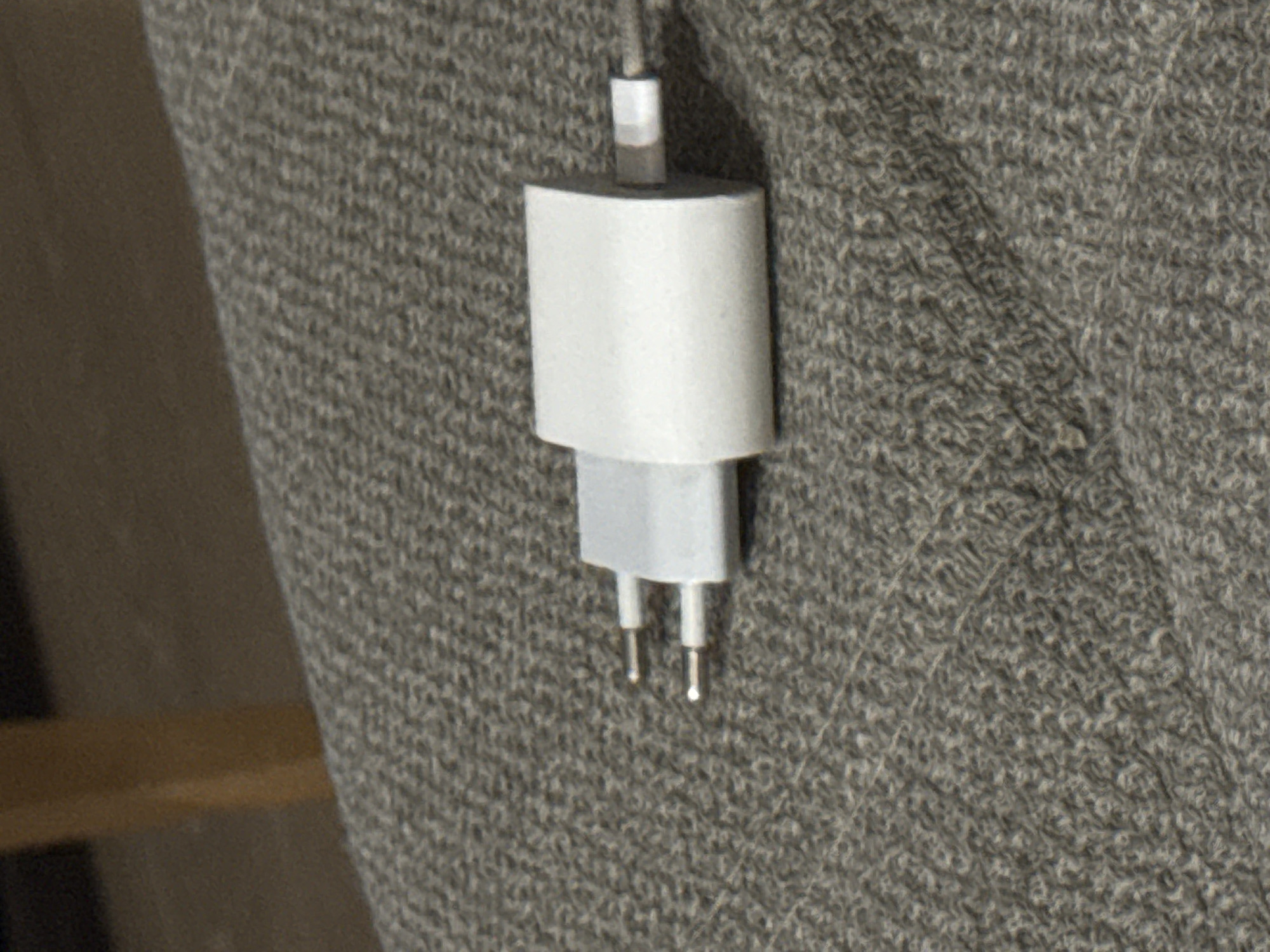 A white iPhone charger is lying on a grey textured surface, possibly a couch or fabric. The charger has two metal prongs on one end and a USB port on the other. The background is blurred and appears to be a wooden surface and a dark wall. There is no indication of the city of Tbilisi, Georgia in the image.