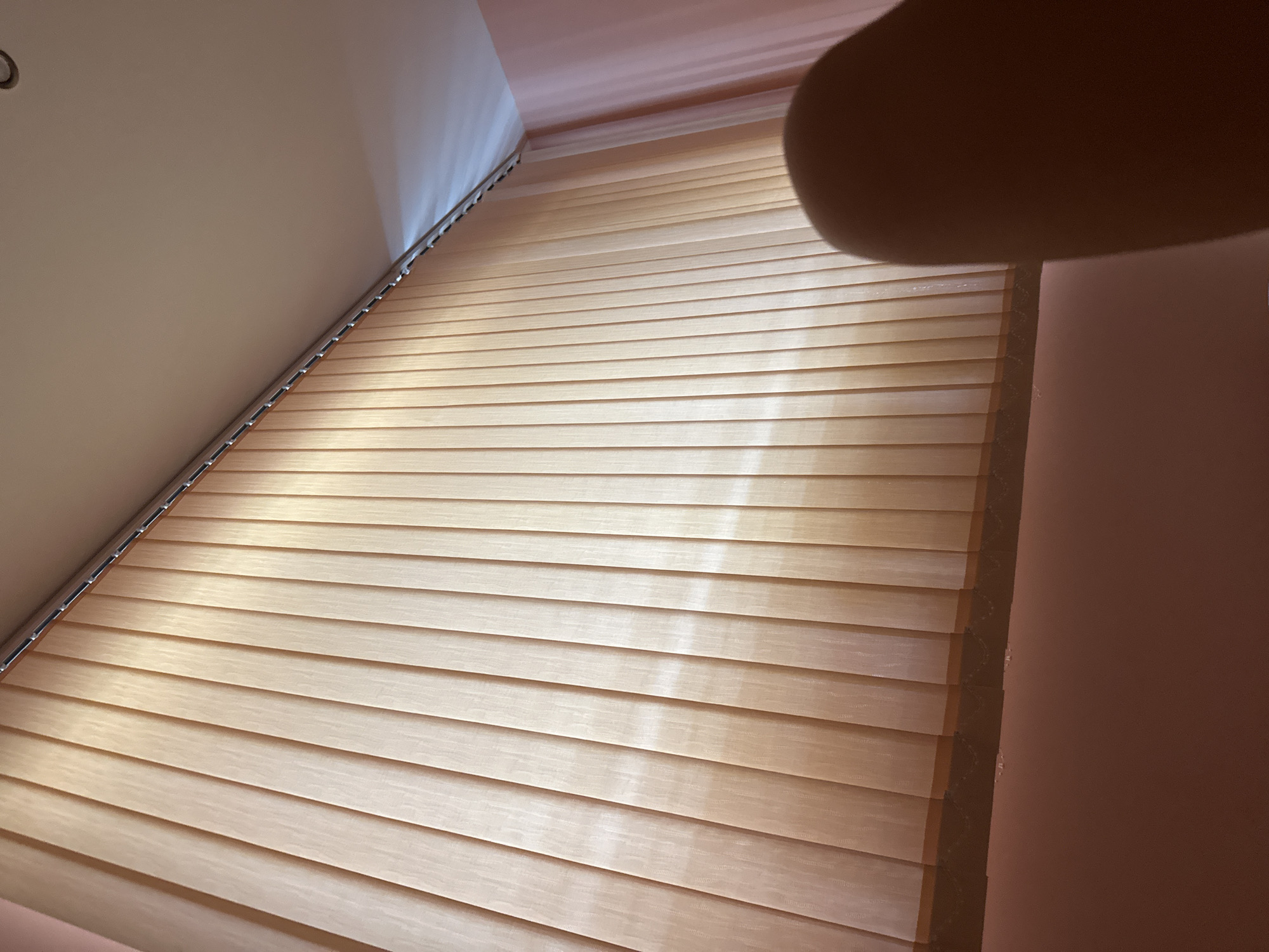 An indoor shot shows a wall of beige vertical blinds that stretch from floor to ceiling. The blinds are illuminated from behind by natural light, creating a soft glow. The ceiling is visible at the top of the image, painted in a light color and featuring a recessed light fixture. A pale pink wall can be seen to the right, with a sheer pink curtain hanging near the corner. In the foreground, a blurred brown object, possibly a person's finger or hand, partially obscures the lower right portion of the blinds. The scene appears to be a room in a residential or commercial building, the specific location of which is unknown.