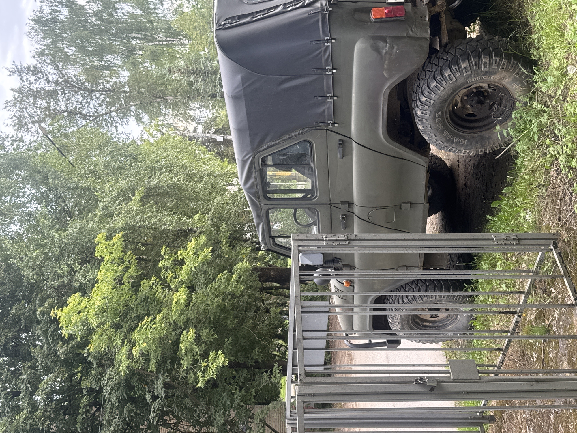 A parked, olive-drab military-style SUV with a black canvas roof is visible in front of a metal gate. The vehicle has large, rugged tires and an exposed spare tire on the rear. The gate is made of vertical metal bars and appears to be closed. The background consists of lush green trees and foliage, with a hint of a building on the left side. The ground is covered with dirt and grass. The scene appears to be in an outdoor urban or suburban setting, possibly a yard or parking area. The image was taken in Dedovsk, Russia. There are no people visible in the image.