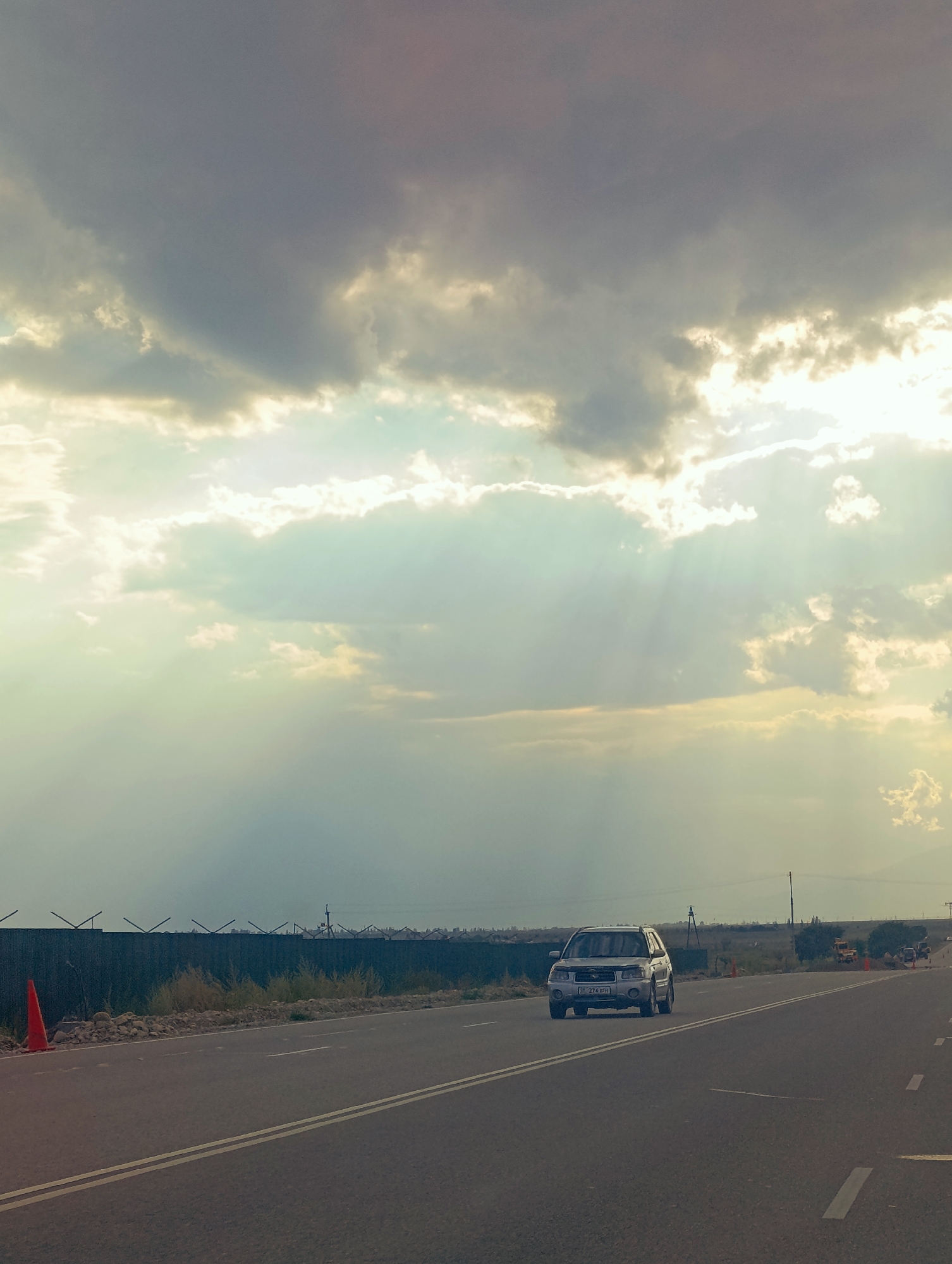 The image depicts a view of a road with a silver SUV driving towards the viewer. The road has a double yellow line on the left side and white dashed lines on the right. In the background, there is a fence with barbed wire, and behind it, dry vegetation and a few trees. The sky is dramatic with heavy clouds, but sunlight is breaking through, creating visible rays of light. There is an orange traffic cone on the left side of the road, near the fence. The scene appears to be in Toru-Aigyr, Kyrgyzstan.