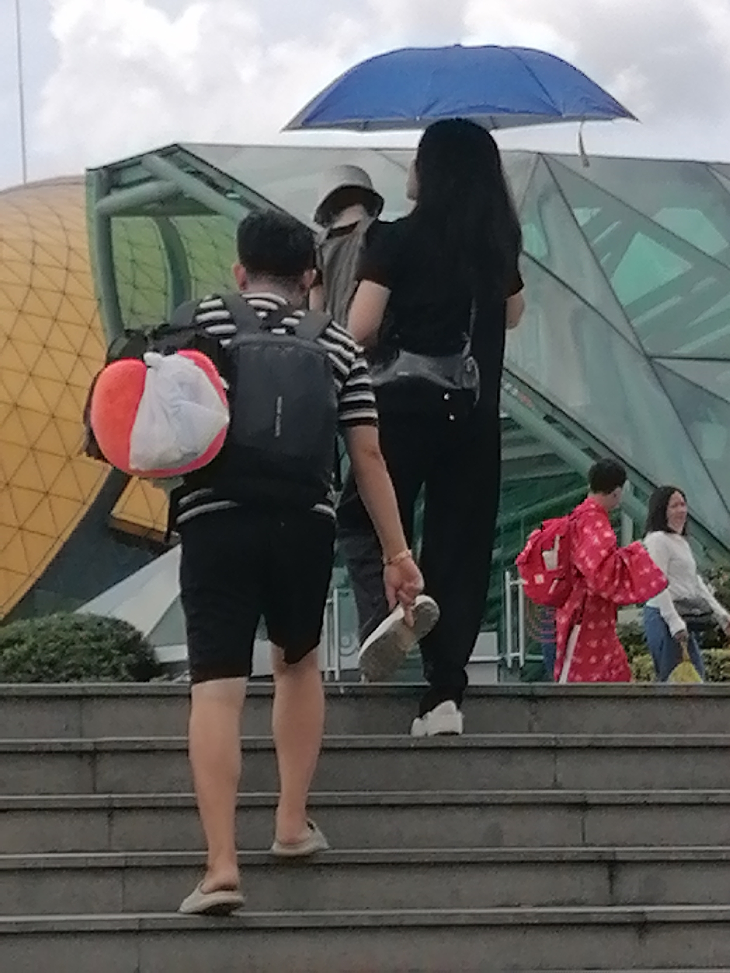 In Da Lat, Vietnam, several individuals ascend a wide set of stone stairs. The scene appears to be near a modern architectural structure with a rounded, golden-colored facade and a large, green, angled glass roof. In the foreground, a person wearing a black and white striped t-shirt, black shorts, and a backpack with a red and white circular object is walking up the stairs. They are also carrying a white plastic bag. To their right, a woman in a black shirt and pants walks under a blue umbrella. Further up the stairs, a person in a red, patterned garment with a red backpack is visible, and to their right, a woman in a white top and blue jeans walks. The sky is overcast with white clouds.