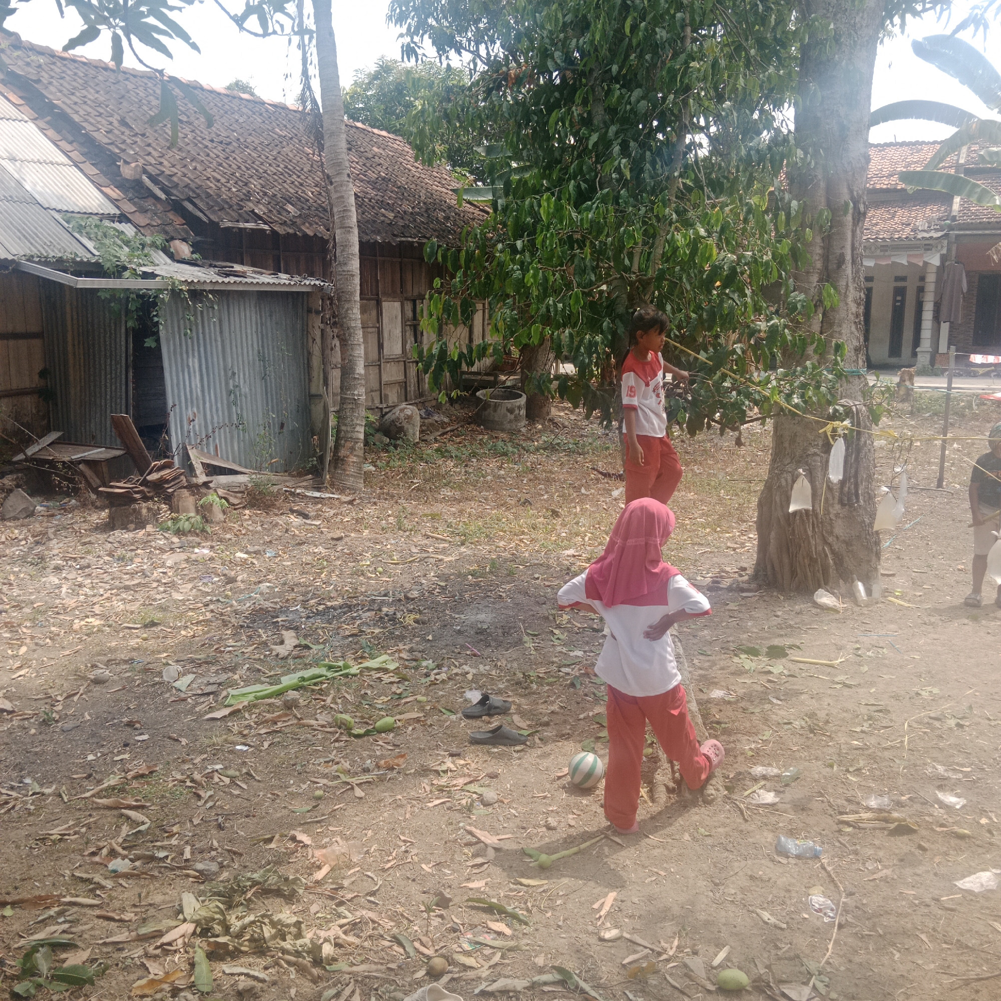 In Tegowanu Kulon, Indonesia, an outdoor scene depicts three individuals in a dusty, debris-strewn yard with a backdrop of modest dwellings. On the left, a dwelling with a tiled roof and corrugated metal siding shows signs of disrepair. To the right, a large tree stands centrally, with a rope strung around it, to which several white plastic bags are attached.
Two children, a boy and a girl, are visible. The girl, wearing a red hijab and a white and red shirt with matching red pants, is positioned in the foreground, facing away from the camera with her hands on her hips. She is next to a striped green and white ball. Further back and to the left, a boy in a white shirt with red trim and red pants stands near a tree. A third figure, a person wearing a dark shirt and light-colored pants, is partially visible on the far right, holding a stick.
The ground is covered in dirt, fallen leaves, twigs, and scattered pieces of debris, including a plastic bottle and discarded footwear. Green leaves and some fruit are also present on the ground. The overall environment suggests a rural or semi-rural residential area.