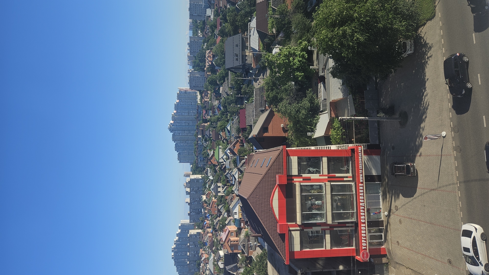 This is an aerial view of a city in Krasnodar, Russia, under a clear blue sky. In the foreground, a red and white commercial building with large display windows is visible. The sign above the entrance reads "ТОРГОВОЕ ОБОРУДОВАНИЕ" which translates to "TRADE EQUIPMENT" and the website "WWW.TO-KRASNODAR.PRO". Several mannequins are displayed inside the windows. In front of the building is a paved parking lot with a few cars, including a dark-colored SUV and a white sedan. A street runs alongside the parking lot, with more vehicles visible.
Beyond the commercial building, the landscape transitions to a dense neighborhood of smaller residential houses with various roof colors. Further in the background, a cluster of tall, multi-story apartment buildings dominates the skyline, indicating a mix of residential and commercial development within the city. Lush green trees are interspersed throughout the scene, adding to the urban greenery. The overall impression is of a sunny day in a moderately developed urban area.