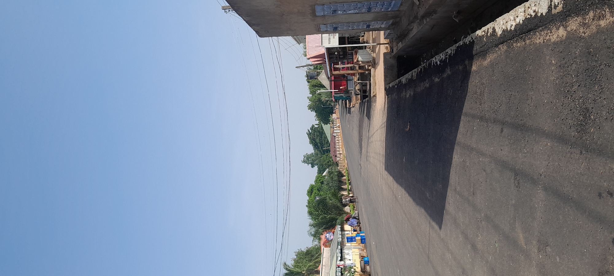A street scene in what appears to be a village or town in Africa under a clear blue sky. The asphalt road curves gently into the distance. On the left side of the road, there are low-lying buildings with awnings, some displaying goods such as blue plastic crates. A few people are visible near these structures, including two men walking away from the camera and another individual seated. Lush green trees and palm trees line the left side of the road. On the right side of the road, there are more structures, including stalls with corrugated metal roofs and a larger building with a rough, plastered exterior. A concrete drainage ditch runs alongside the road on the right. Power lines are visible stretching across the sky. The scene suggests a daytime setting with natural light.