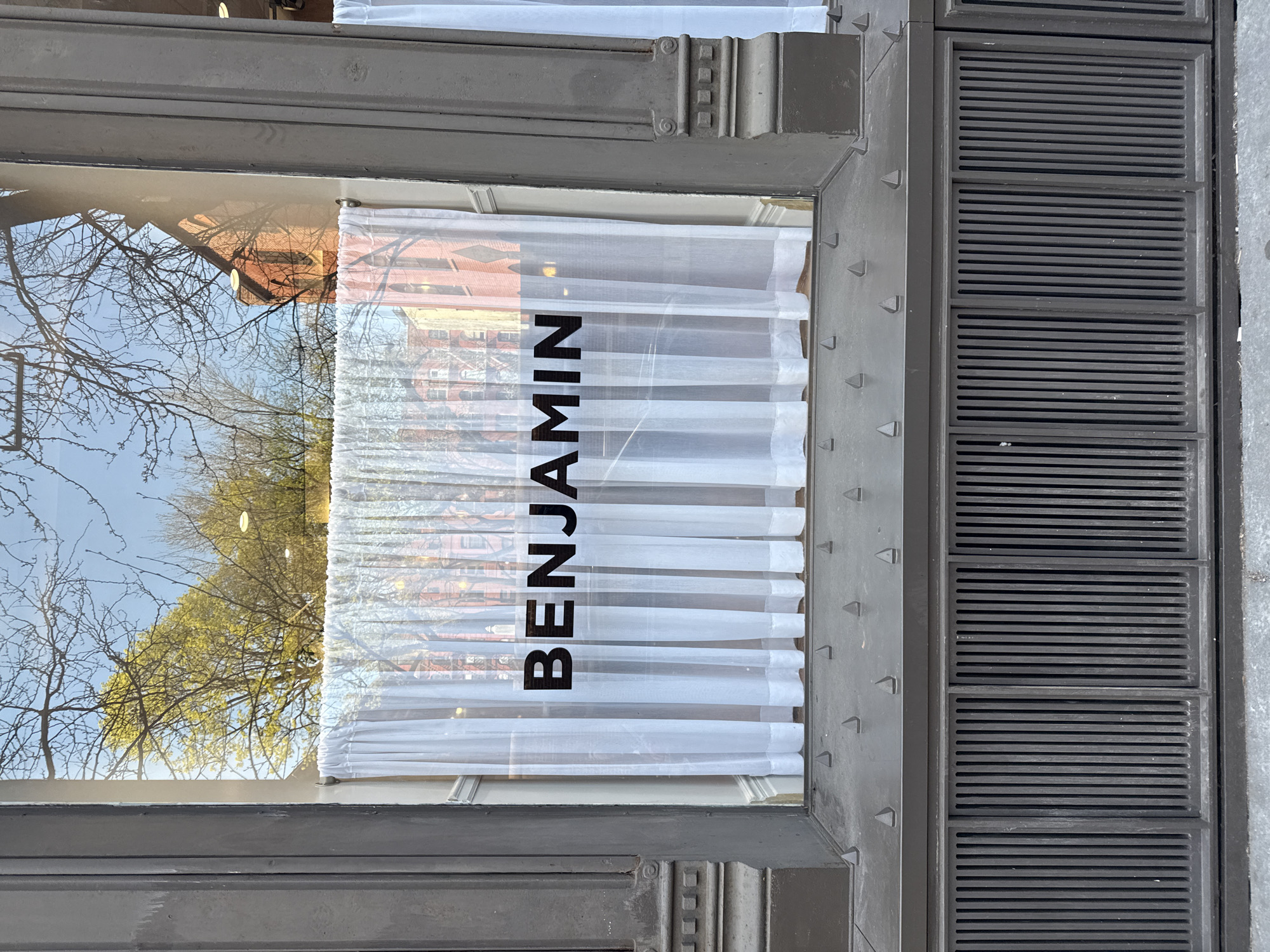 The image shows a storefront window with the name "BENJAMIN" vertically printed in black on a white translucent curtain. The reflection in the window shows bare trees and a brick building, suggesting a neighborhood setting. The window frame is a dark color and has ornate details. Adjacent to the window is a ventilation grate with horizontal slats. Based on the architecture and design details, it is likely located in the city of New York, United States of America.