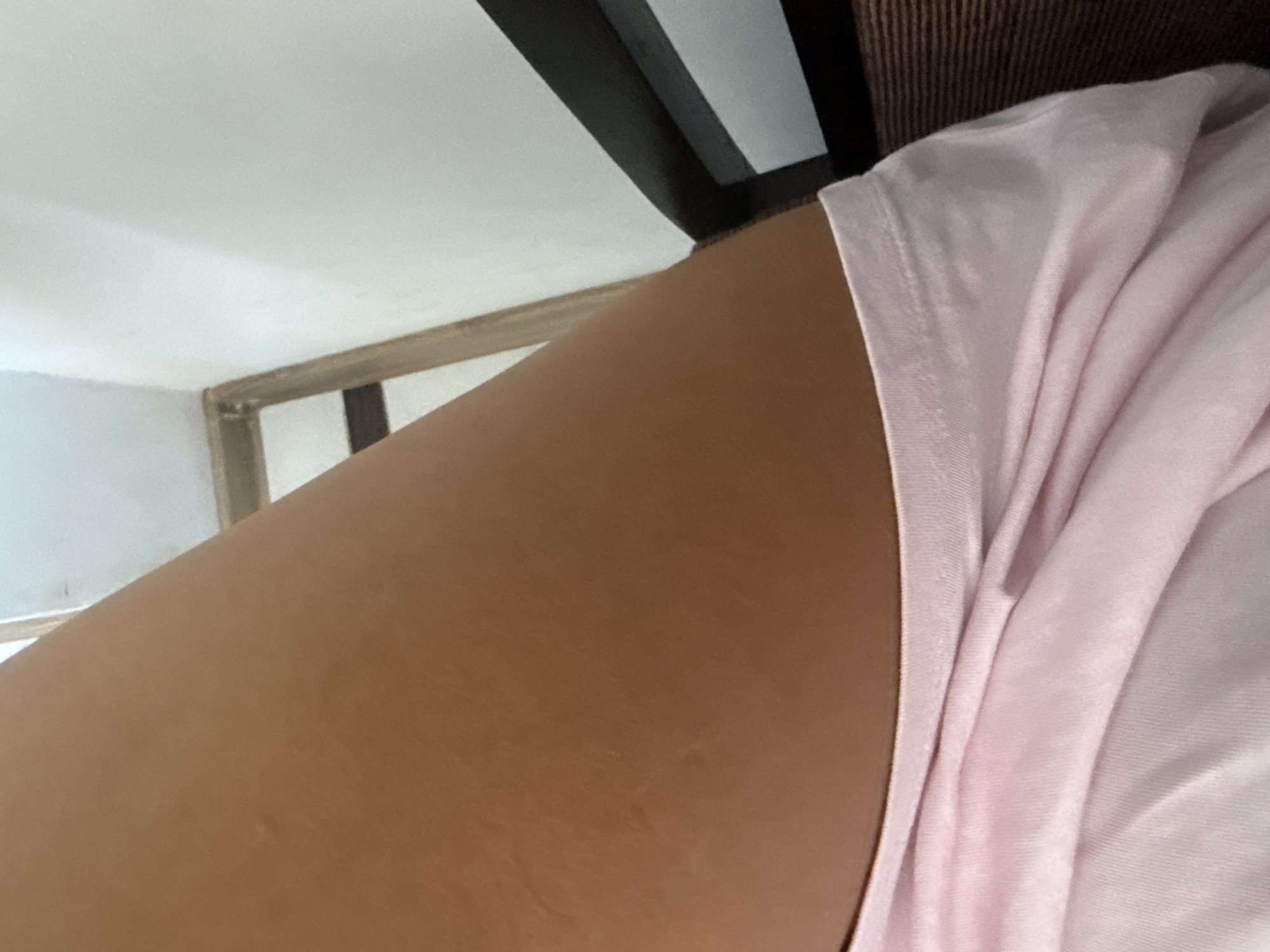 A close-up, angled shot focuses on a person's tanned leg and the top edge of a pink garment, likely a t-shirt. The leg exhibits a few subtle marks or indentations on the skin. The pink fabric is draped and creased, creating soft shadows and highlights. In the background, a white wall meets a wooden frame and a dark-colored object, possibly furniture. The setting appears to be indoors, with neutral lighting. The provided context of Chișinău, Moldova, offers no direct visual cues within the image itself, as the scene is intimate and lacks distinguishing geographical features.