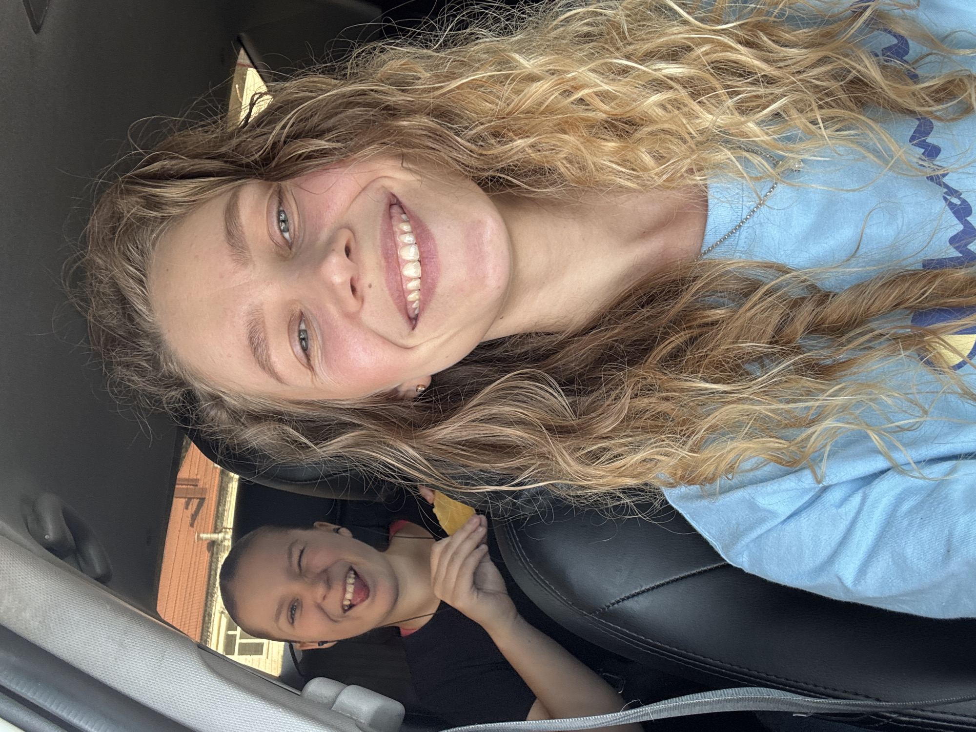 Two smiling females are in a car in Kurganinsk, Russia. One is the foreground with blonde curly hair and a blue t-shirt, while the other is in the back. The younger looking person in the back is in a black t-shirt and is holding a yellow rectangular item. A black leather car seat can be seen behind the background person. The car is stationary in front of brick buildings.