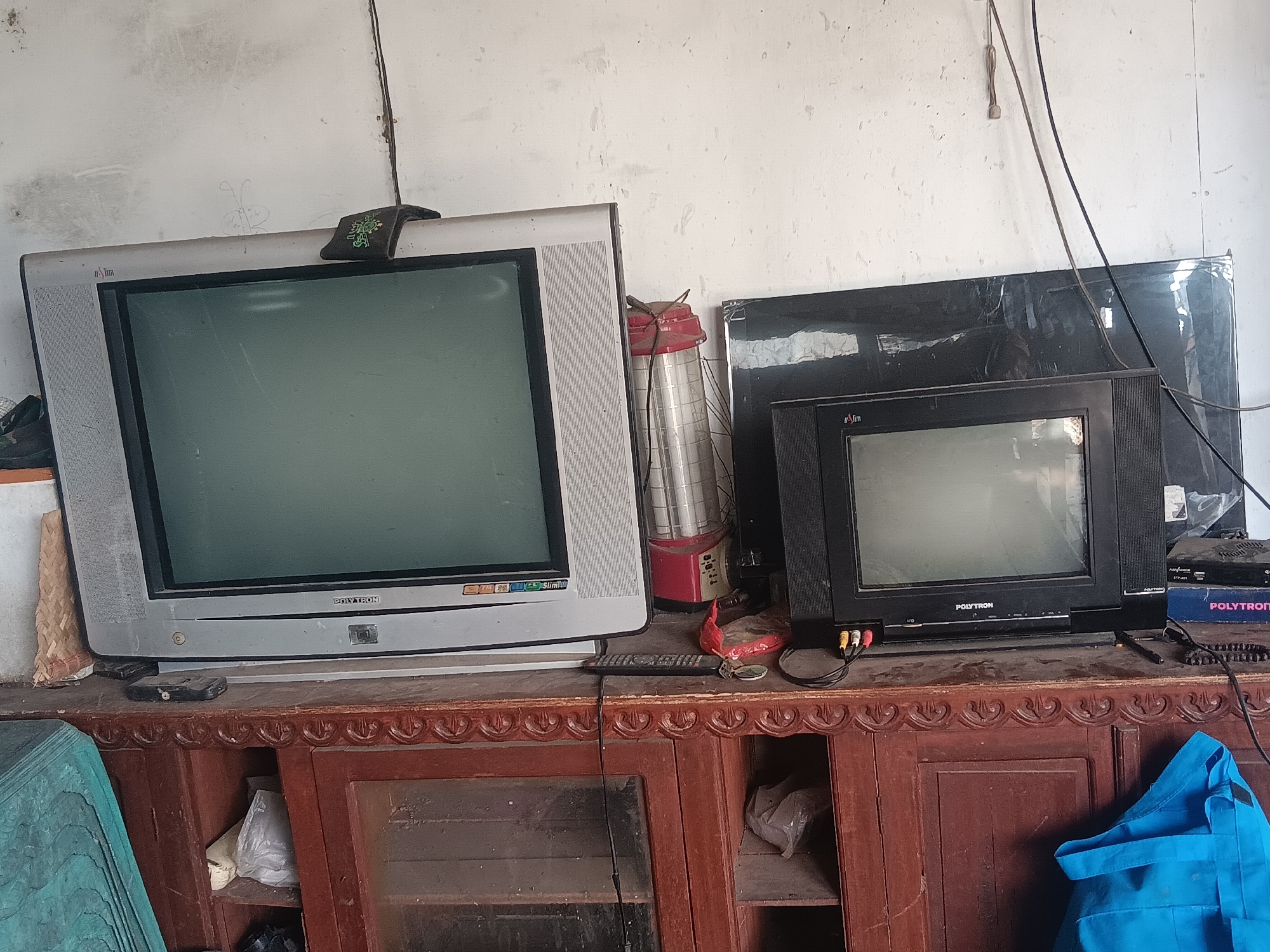 In Batursari, Indonesia, two old television sets are positioned on a wooden cabinet. The television on the left is a silver, bulky CRT model with a green screen. A small, black object with a green logo is placed on top of it. To the right of this television is a red and white lantern. Next to the lantern, a smaller, black CRT television with a grey screen is situated. It has "POLYTRON" written on its front. The televisions and other objects rest on a dark brown wooden cabinet with decorative carvings along the top edge and glass doors. Various cables and a blue bag are also visible. The overall environment appears to be indoors, with a plain, textured wall in the background.