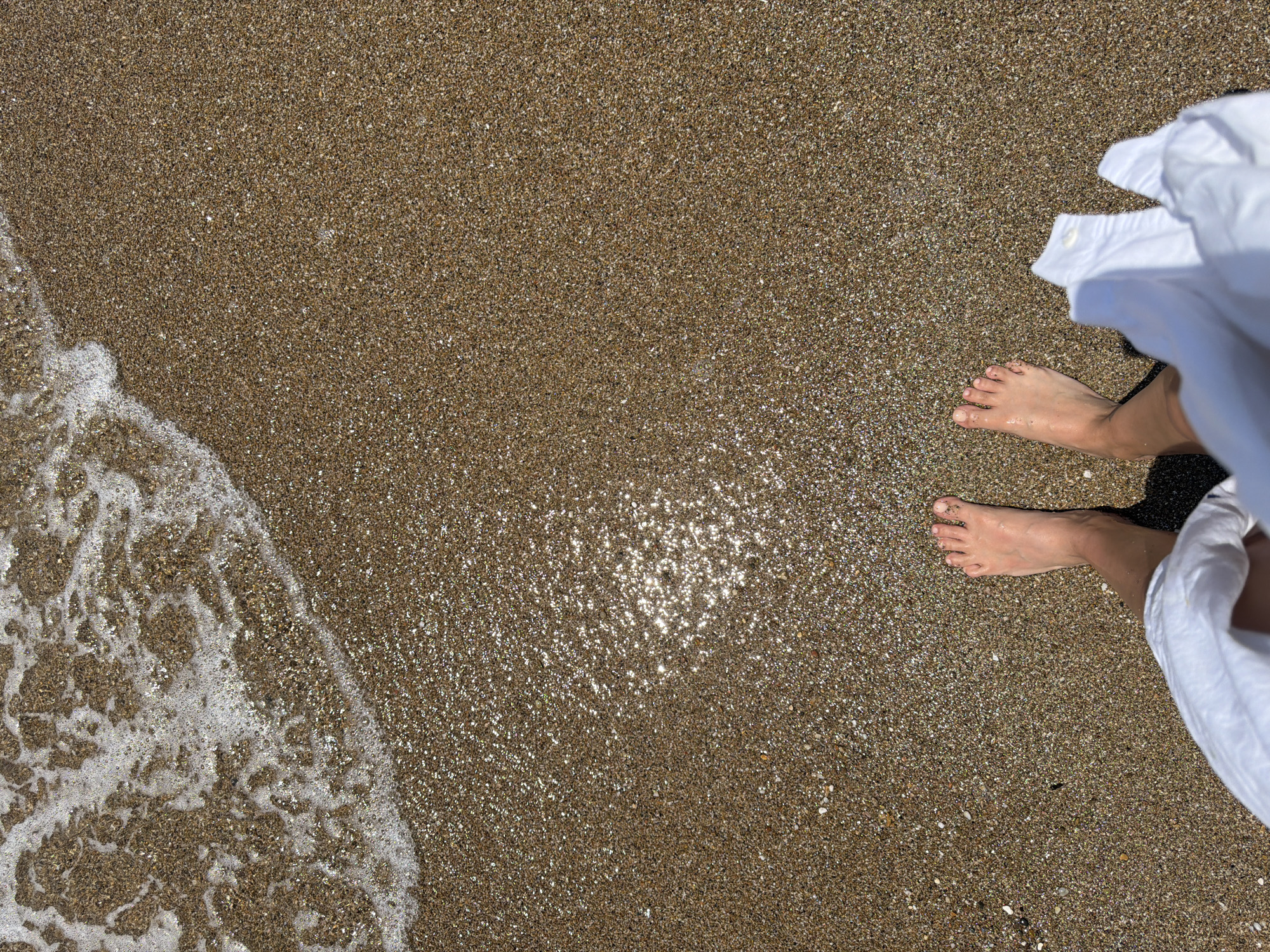 An overhead shot captures a person's bare feet standing on a shimmering wet beach at the water's edge on a bright, sunny day, likely in Νέα Χηλή, Greece. The beach surface is composed of fine, varied-colored pebbles and sand, predominantly in shades of brown and gold, which glisten intensely with reflected sunlight, particularly noticeable in the upper-middle and lower-right areas of the frame.
On the bottom-left, a foamy white wave, either receding or gently washing ashore, creates a dynamic, curved line across the sand. The person's bare feet are positioned in the lower-right quadrant, with the right foot more prominently displayed. The left foot is partially visible behind it. Part of a loose, white garment, possibly a shirt or dress, is visible on the far right side of the frame, billowing slightly. A dark shadow, likely cast by the person, falls behind their feet and beneath the white fabric.
The scene evokes a sense of relaxation and connection with nature, suggesting a leisurely moment spent enjoying the sun and the sea. There is no visible text in the image.