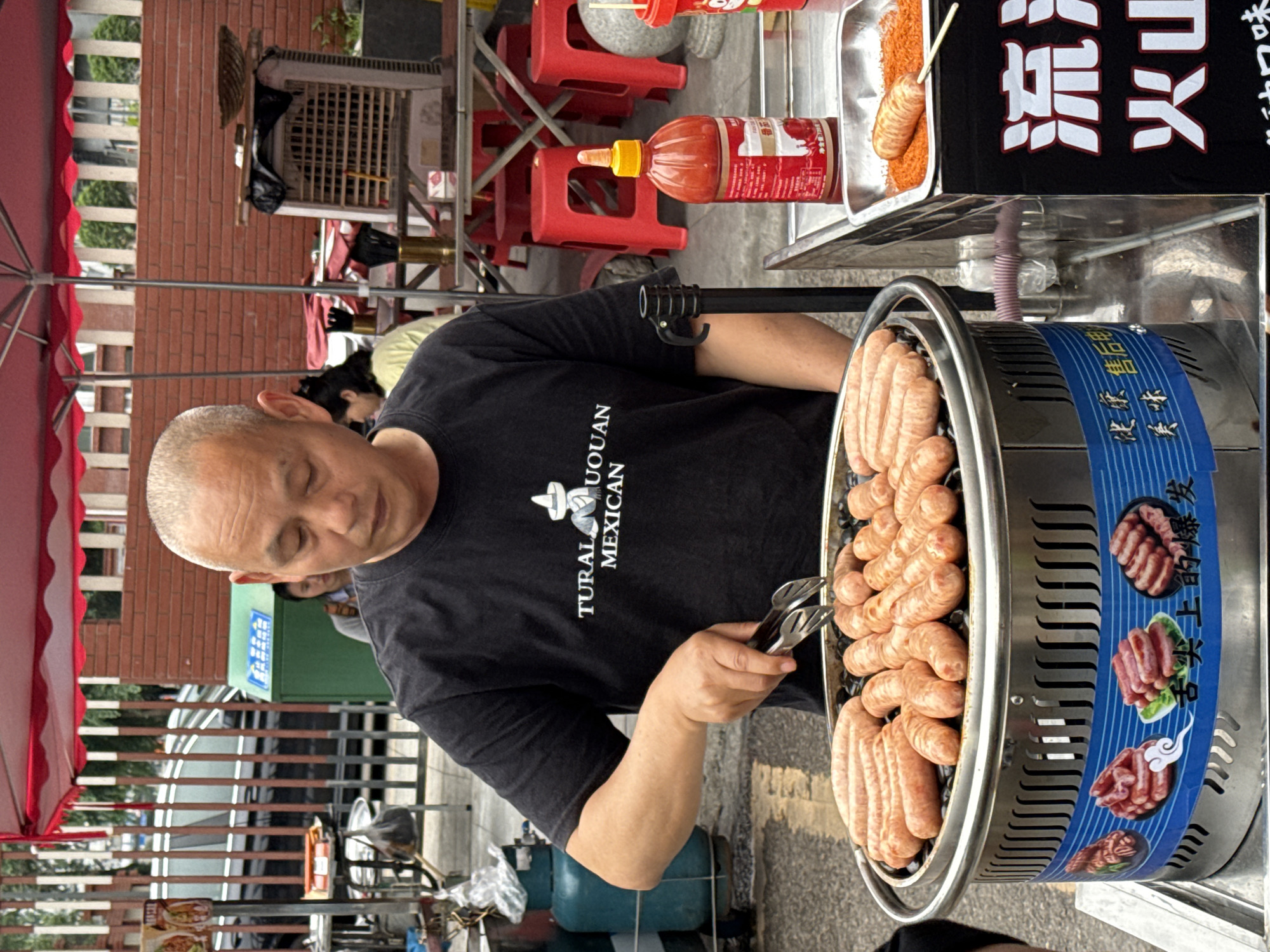 A street food vendor is grilling sausages in Nan'an District, Chongqing, China. The man, with a shaved head and wearing a black T-shirt, is using tongs to turn the sausages on a cylindrical grill. The sausages are arranged in a circular pattern within the grill. Next to the grill is a metal tray with various sauces and a sign in Chinese characters. In the background are red plastic chairs, a brick building, a metal fence, and a glimpse of parked vehicles. A red umbrella is partially visible on the left side of the frame.