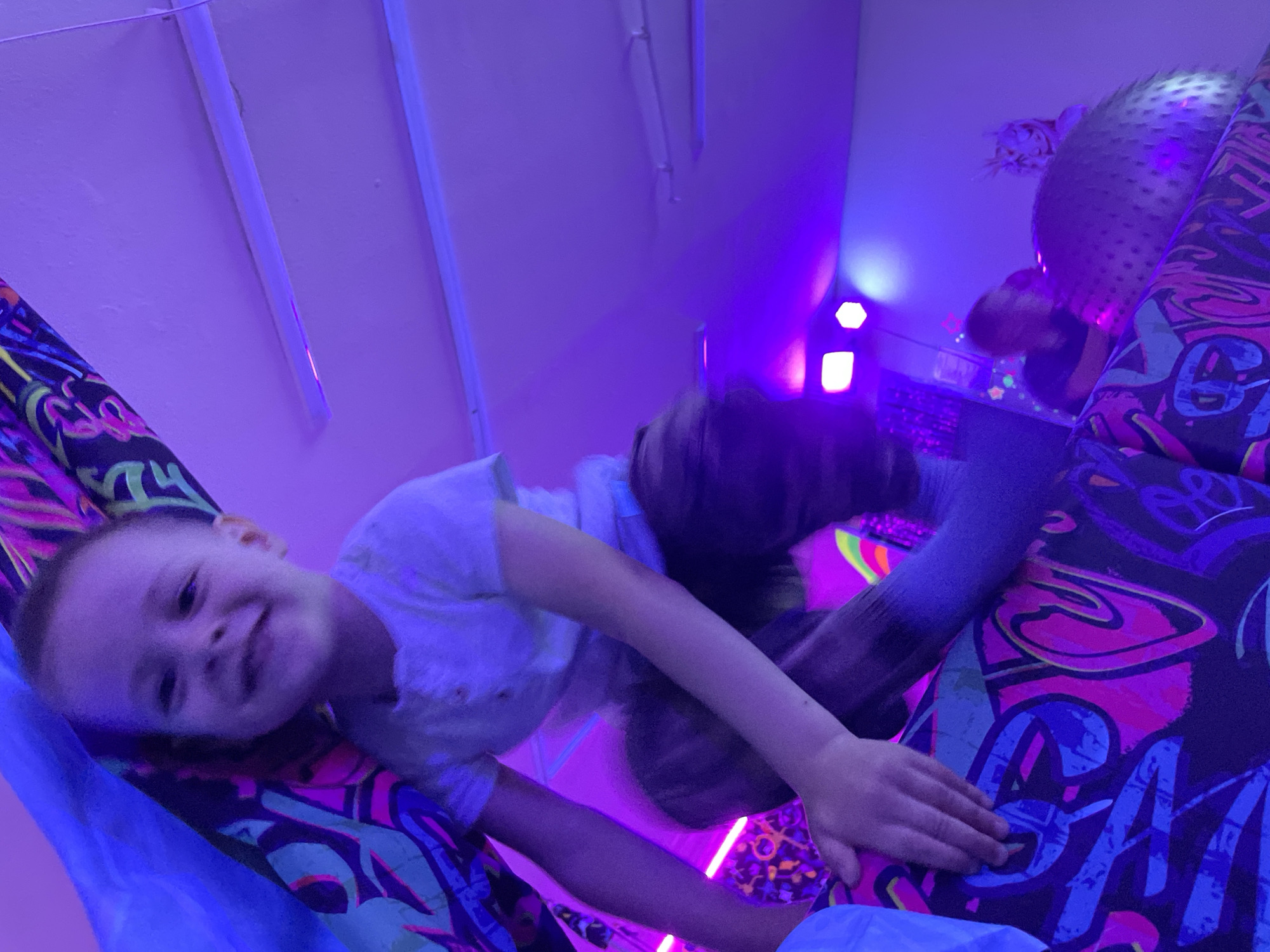The image shows a child lying on a patterned surface under purple lighting. The surface has a mix of black, pink, blue, and green patterns. The child has short hair, a light-colored shirt, and brown pants. The background includes a white ceiling with fluorescent lights, and other items with reflective surfaces. The location is Unknown, Unknown.