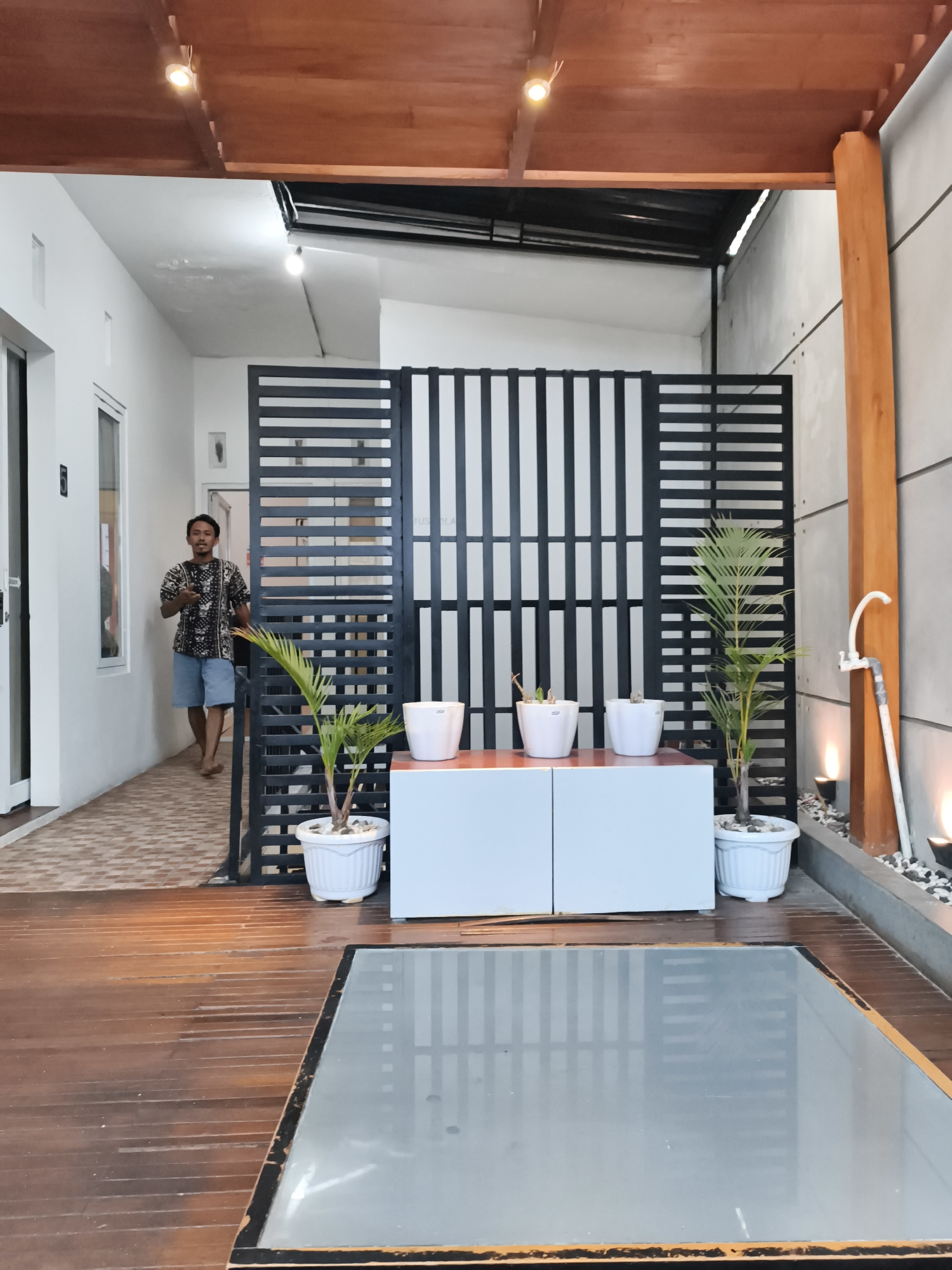 A man in a patterned short-sleeved shirt and blue shorts walks down a tiled hallway towards the viewer, holding a dark object in his right hand. To his right, a black metal screen divides the hallway from an outdoor area with wooden decking. In the outdoor area, a white cabinet holds three potted plants. Further to the right, a wooden post supports a gray wall, with a white faucet and pipe attached to the wall. The scene is illuminated by warm-toned lights from the ceiling and the outdoor area. The overall impression is of a modern outdoor living space adjacent to an indoor hallway. The location is identified as Dieng Kulon, Indonesia.